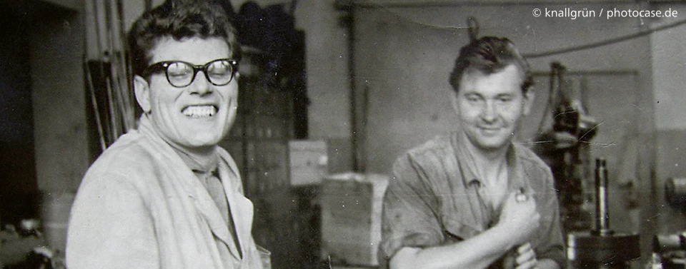 Ein schwarz-weißes Foto zeigt zwei lachende Männer in einer Werkstatt. Einer trägt eine Brille und einen Kittel, der andere ein Arbeitshemd.