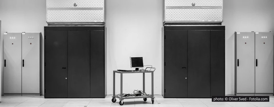 Ein schwarz-weißes Foto eines Serverraums mit zwei großen Serverschränken, Schaltschränken an den Seiten und einem mobilen Wagen mit Monitor und Kabeln in der Mitte.