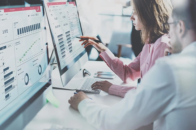 Zwei Personen analysieren Daten auf großen Monitoren in einem modernen Büro. Digitale Dashboards und datengetriebene Entscheidungen stehen im Fokus.