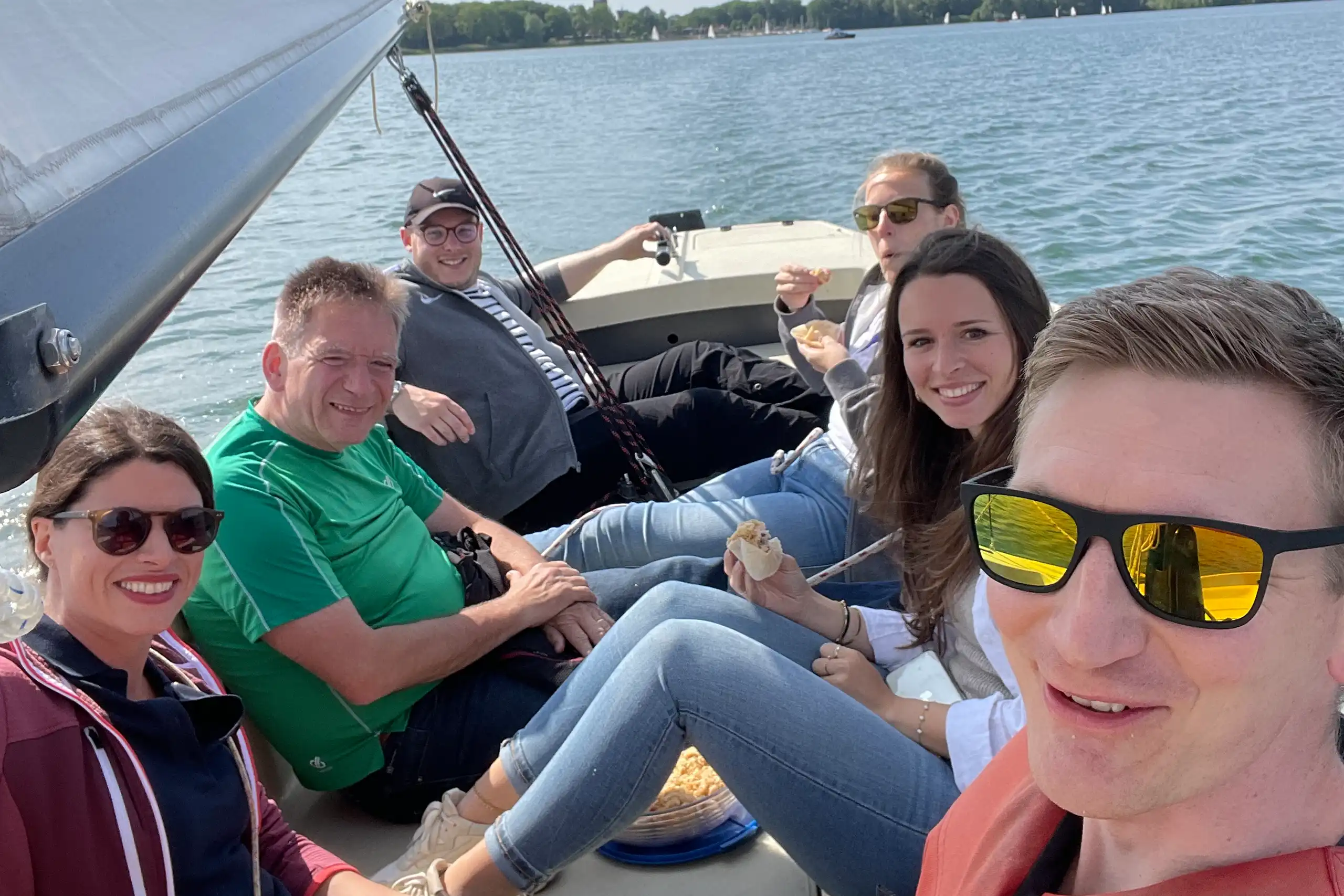 Das Bild zeigt eine Gruppe von sechs Novis, die entspannt auf einem Segelboot auf einem See sitzen, essen und lachen. Im Hintergrund ist Wasser und Uferlandschaft zu sehen.