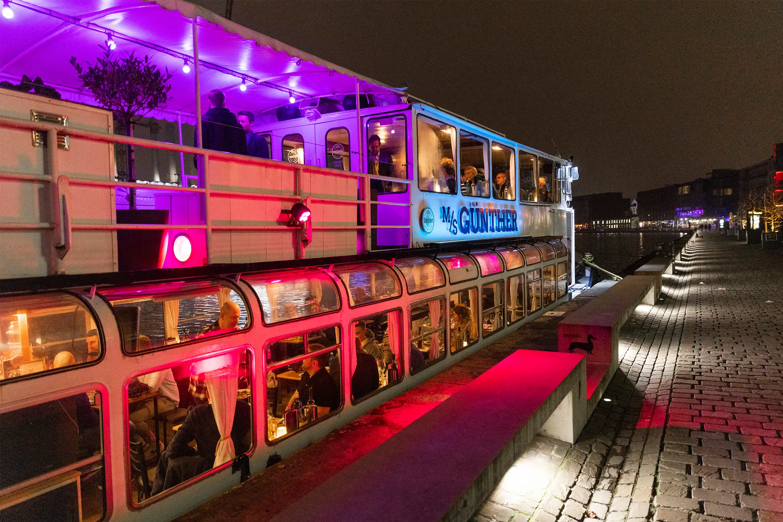 Das Bild zeigt das festliegende Schiff „MS Günther“ bei Nacht, beleuchtet mit buntem Licht. Gäste sitzen im Inneren und auf dem Deck, während das Ufer erleuchtet ist.