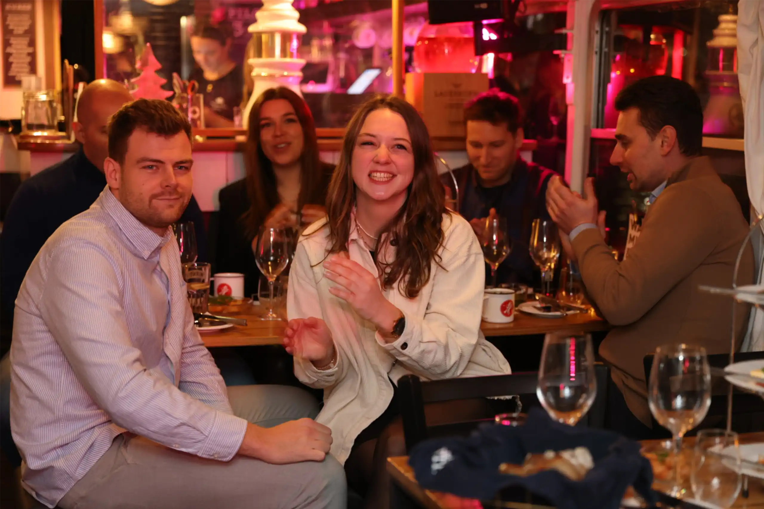 Mehrere Novis sitzen in einer gemütlichen, beleuchteten Umgebung an einem Tisch mit Weingläsern. Eine Frau lacht herzlich, während andere im Hintergrund klatschen.