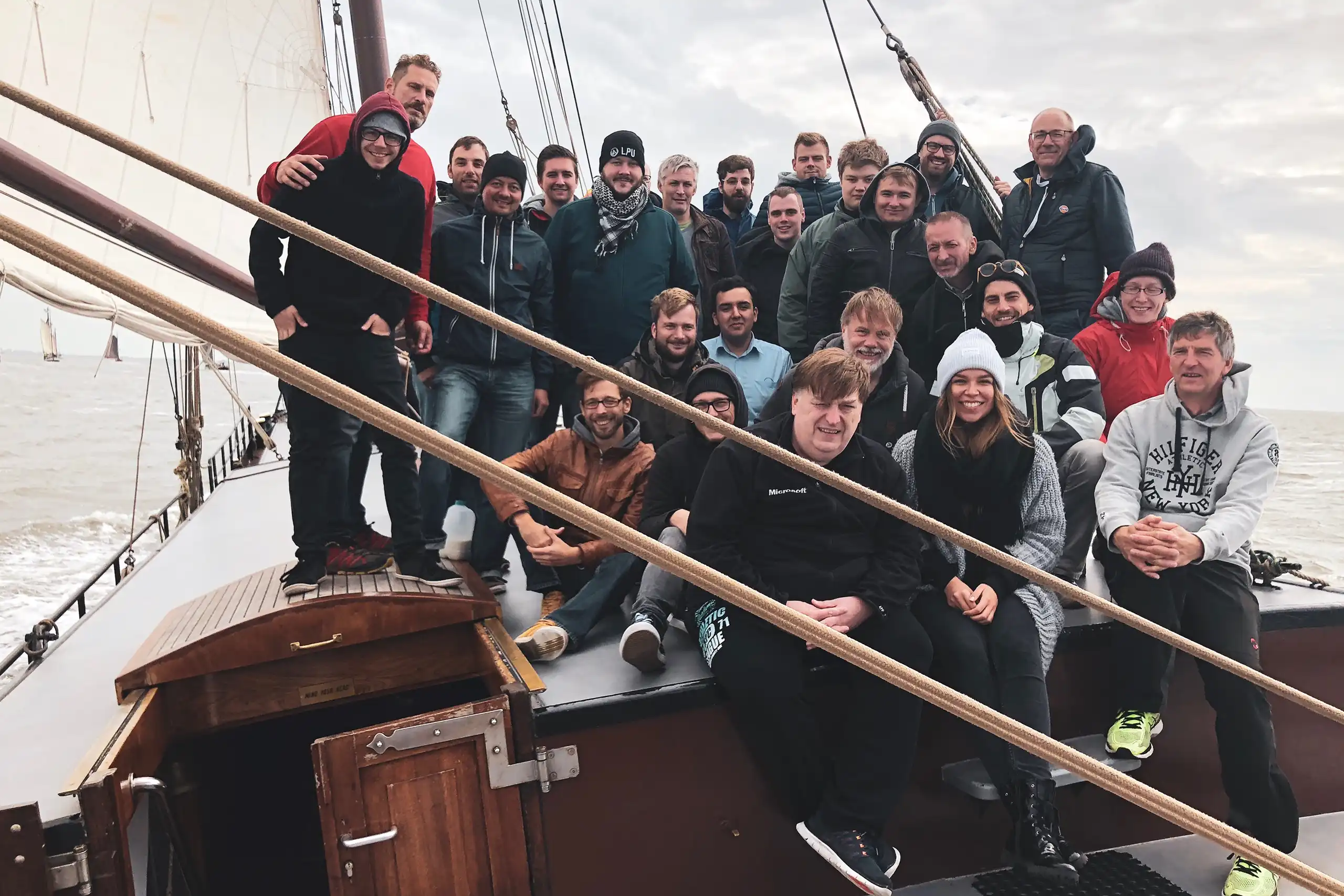 Eine Gruppe von noventum Mitarbeitenden sitzt und steht an Deck eines Segelschiffs auf dem Meer. Sie tragen warme Kleidung und lächeln in die Kamera.