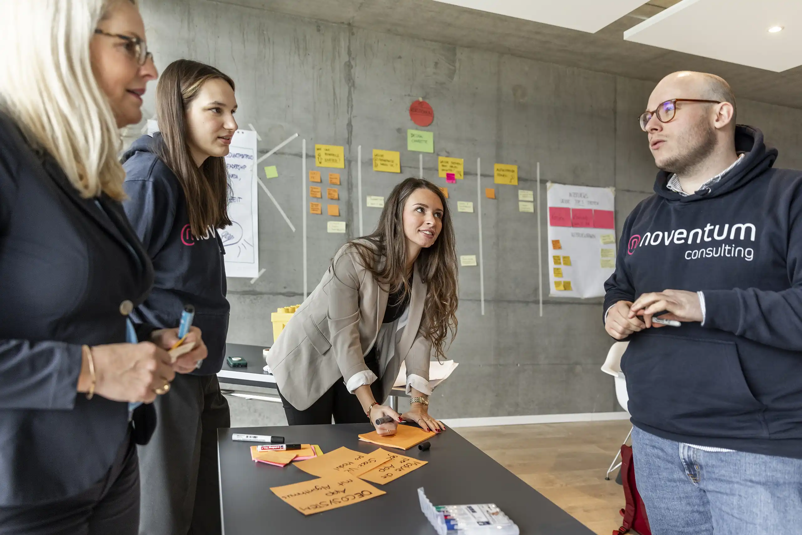 Vier Personen stehen an einem Tisch in einem Workshop. Sie diskutieren und schreiben auf Moderationskarten. Im Hintergrund sind Pinnwände mit Notizen zu sehen.