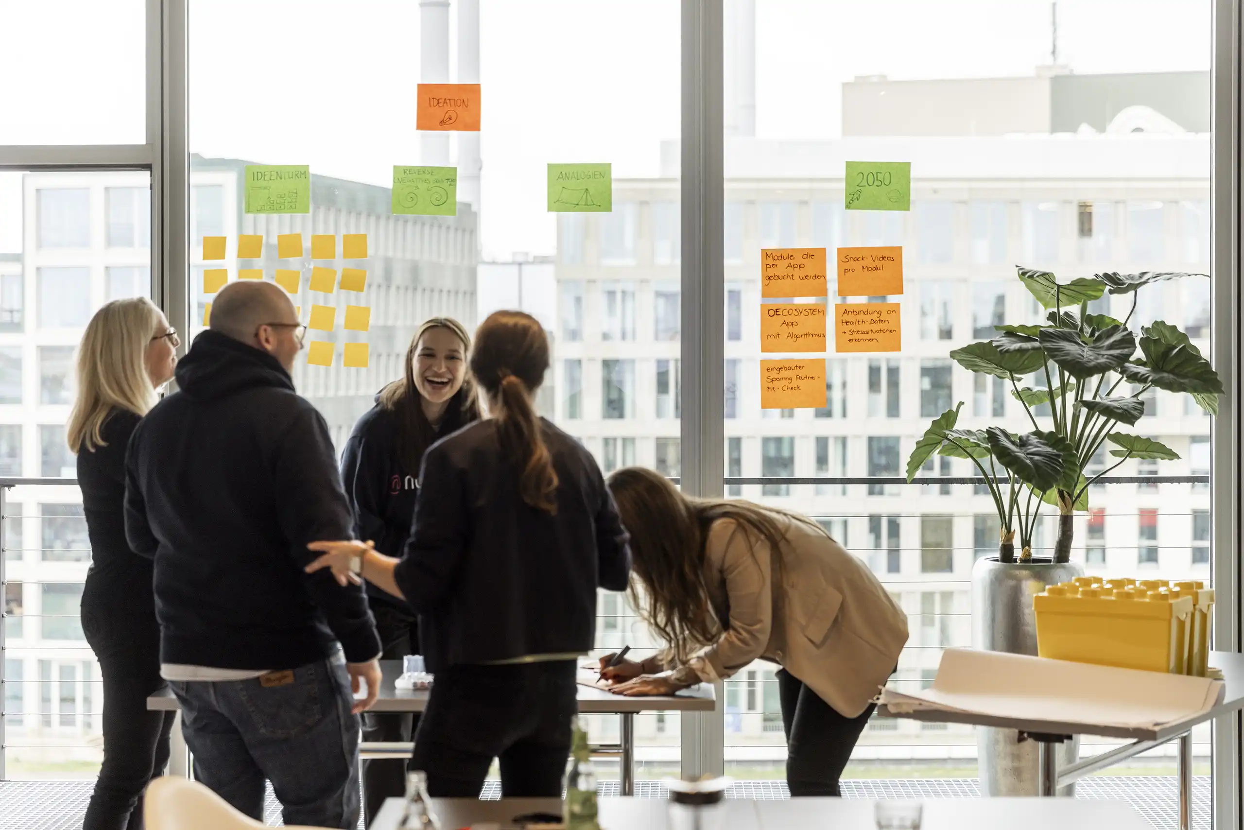 Ein kreativer Workshop mit mehreren Personen, die an einem Tisch arbeiten, Ideen aufschreiben und an Fenstern mit Post-its eine Ideensammlung gestalten.