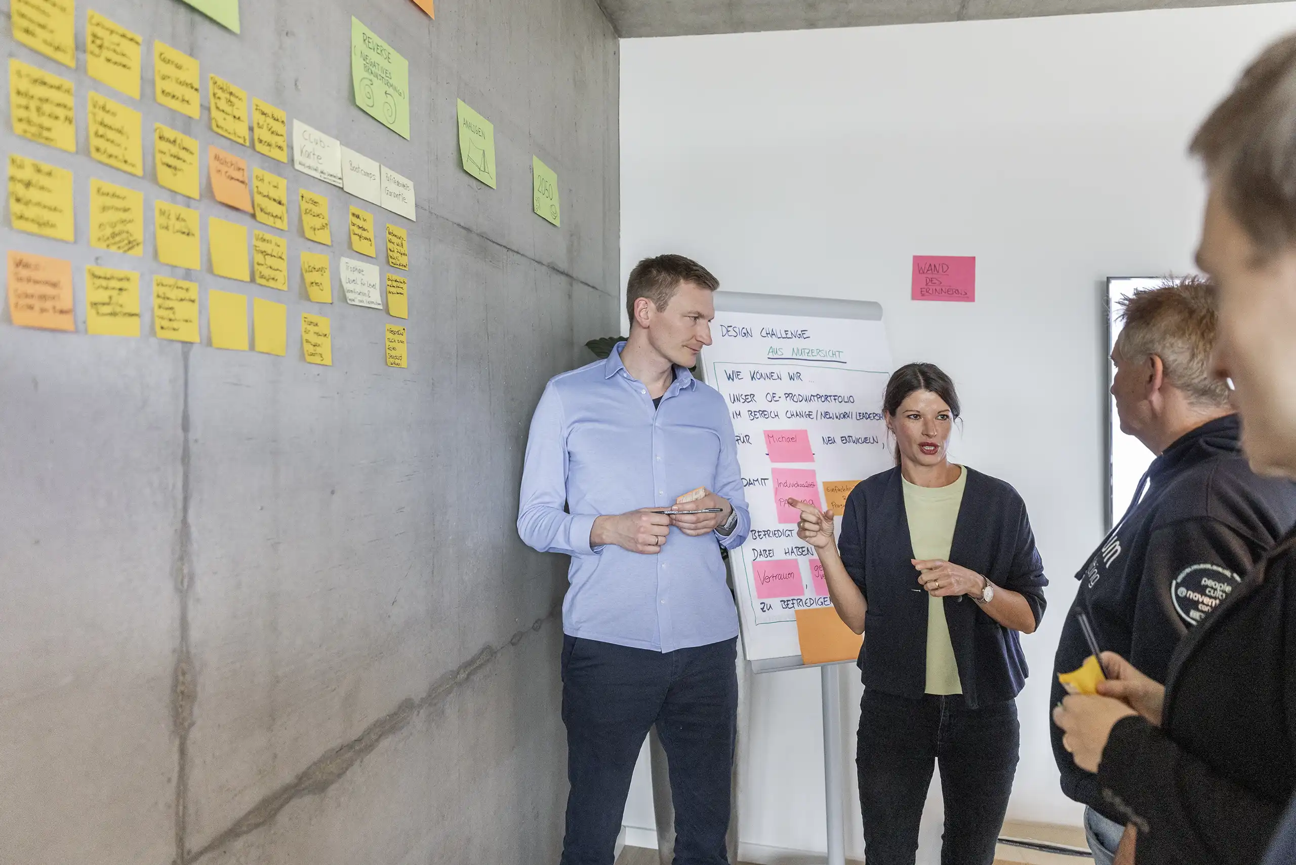 Ein Team steht vor einer Wand mit bunten Notizen und diskutiert Ideen. Eine Frau erklärt etwas anhand eines Flipcharts, während die anderen aufmerksam zuhören.