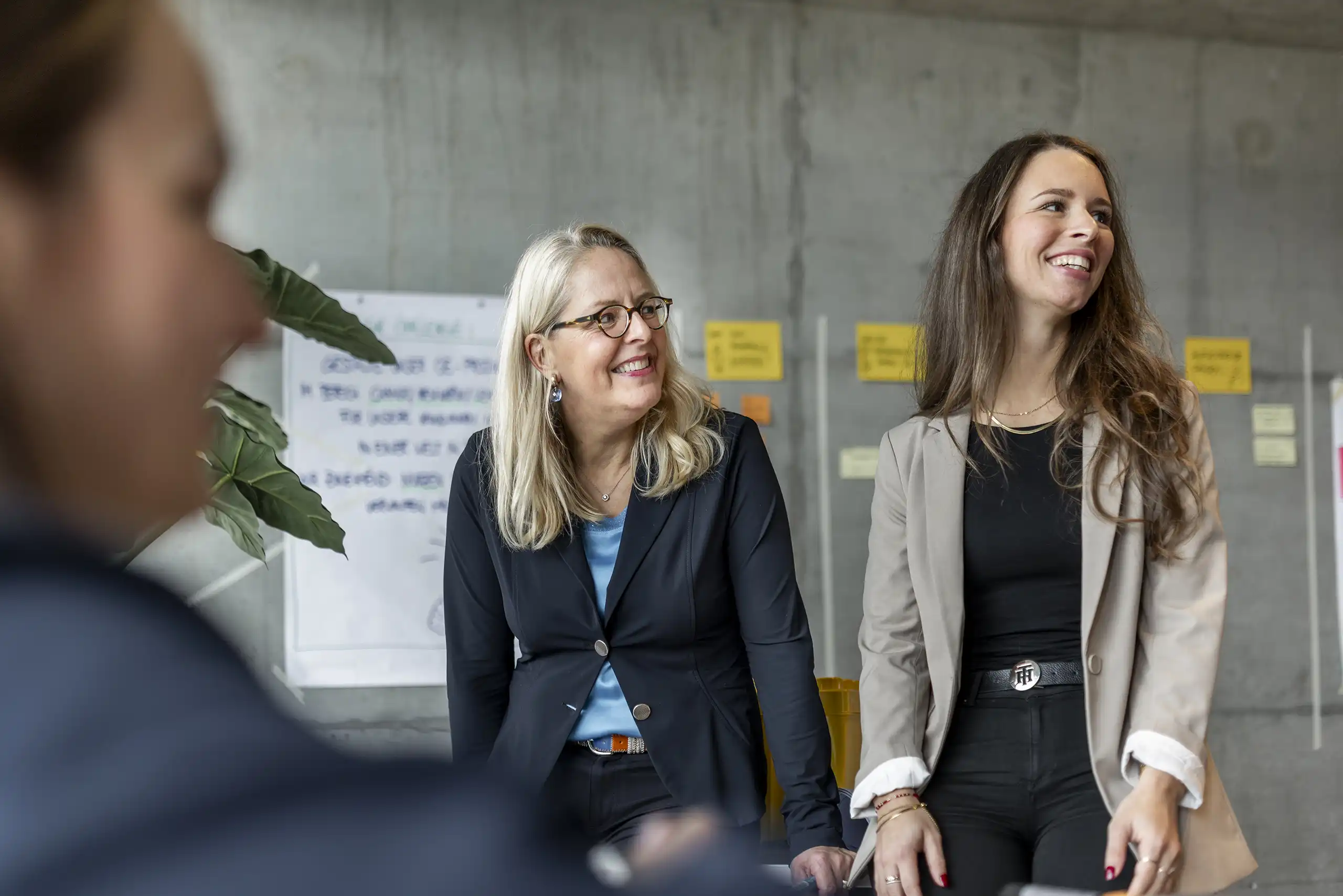 Zwei Frauen stehen lächelnd in einem kreativen Arbeitsumfeld mit Notizen an der Wand, während eine weitere Person im Vordergrund unscharf zu sehen ist.