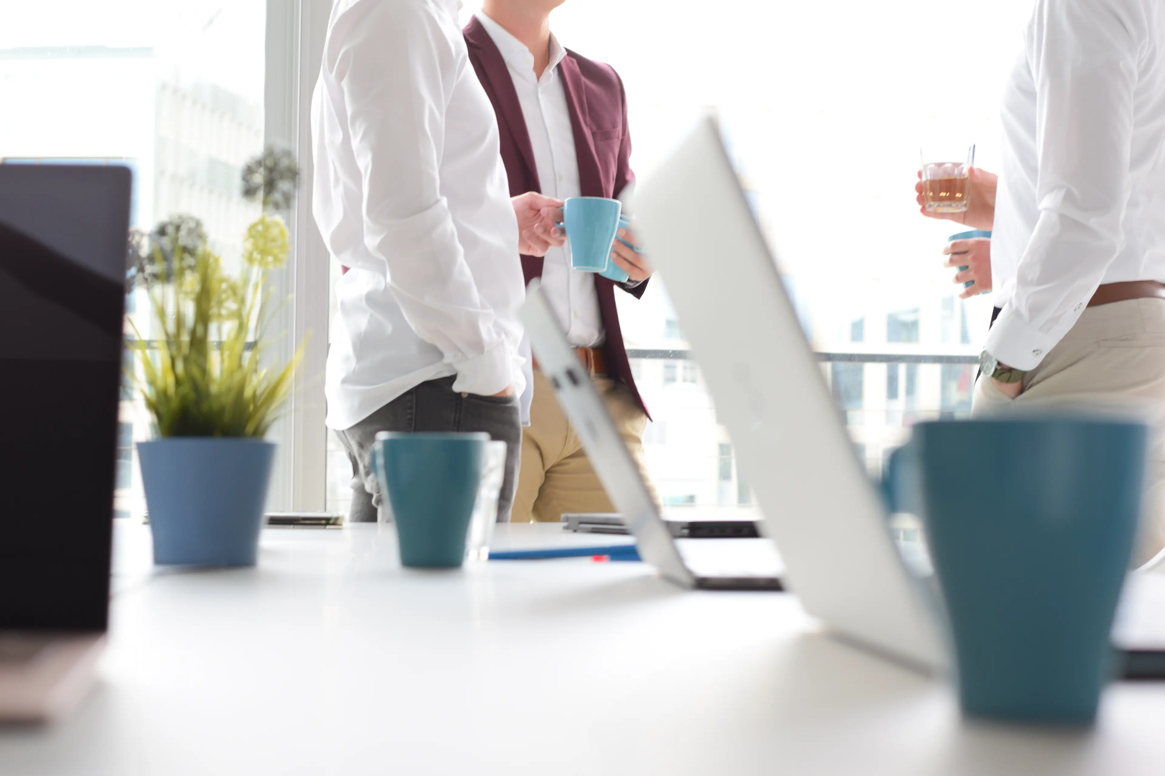 Mehrere Personen stehen in einem modernen Büro, halten Kaffeetassen und unterhalten sich. Im Vordergrund sind Laptops, Pflanzen und Tassen auf einem Tisch.