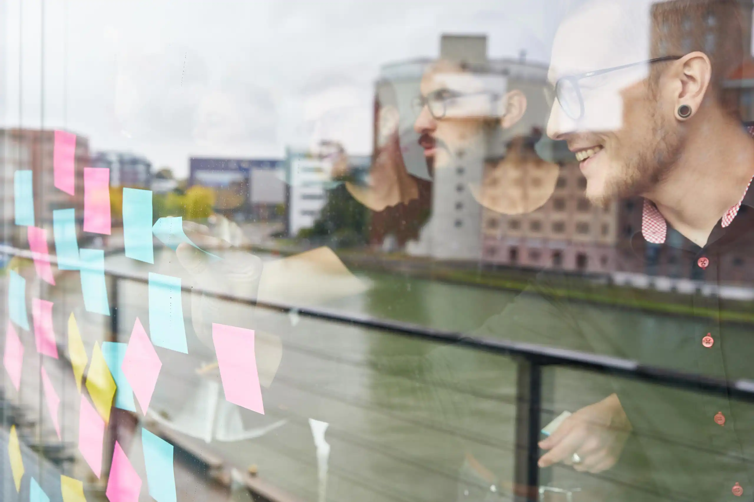 Drei Personen brainstormen an einer Glaswand mit bunten Haftnotizen. In der Glasscheibe spiegelt sich die Stadt.