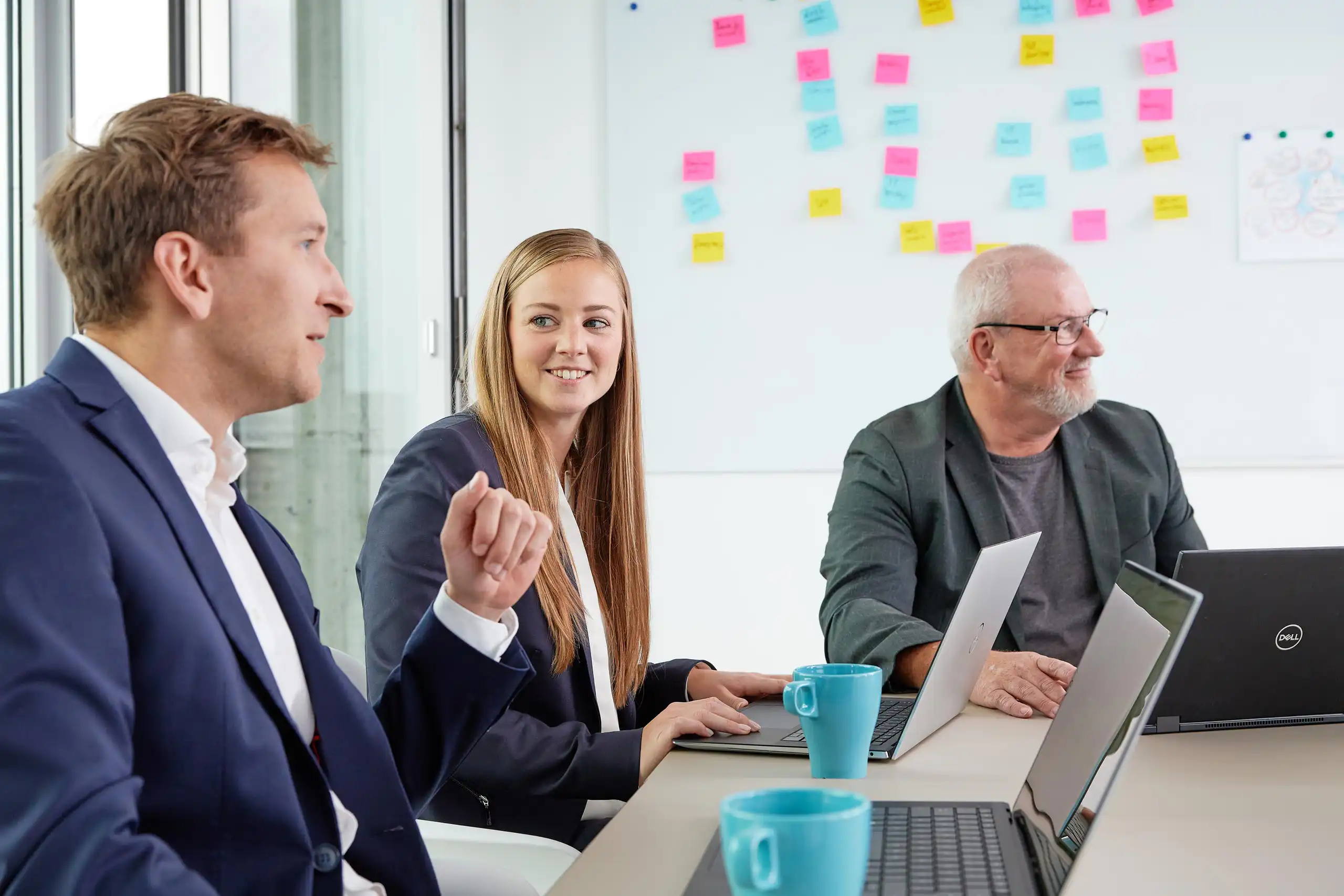 Drei Personen in Business-Kleidung sitzen an einem Tisch mit Laptops und Tassen, diskutieren und lächeln. Im Hintergrund eine Whiteboard-Wand mit bunten Notizen.