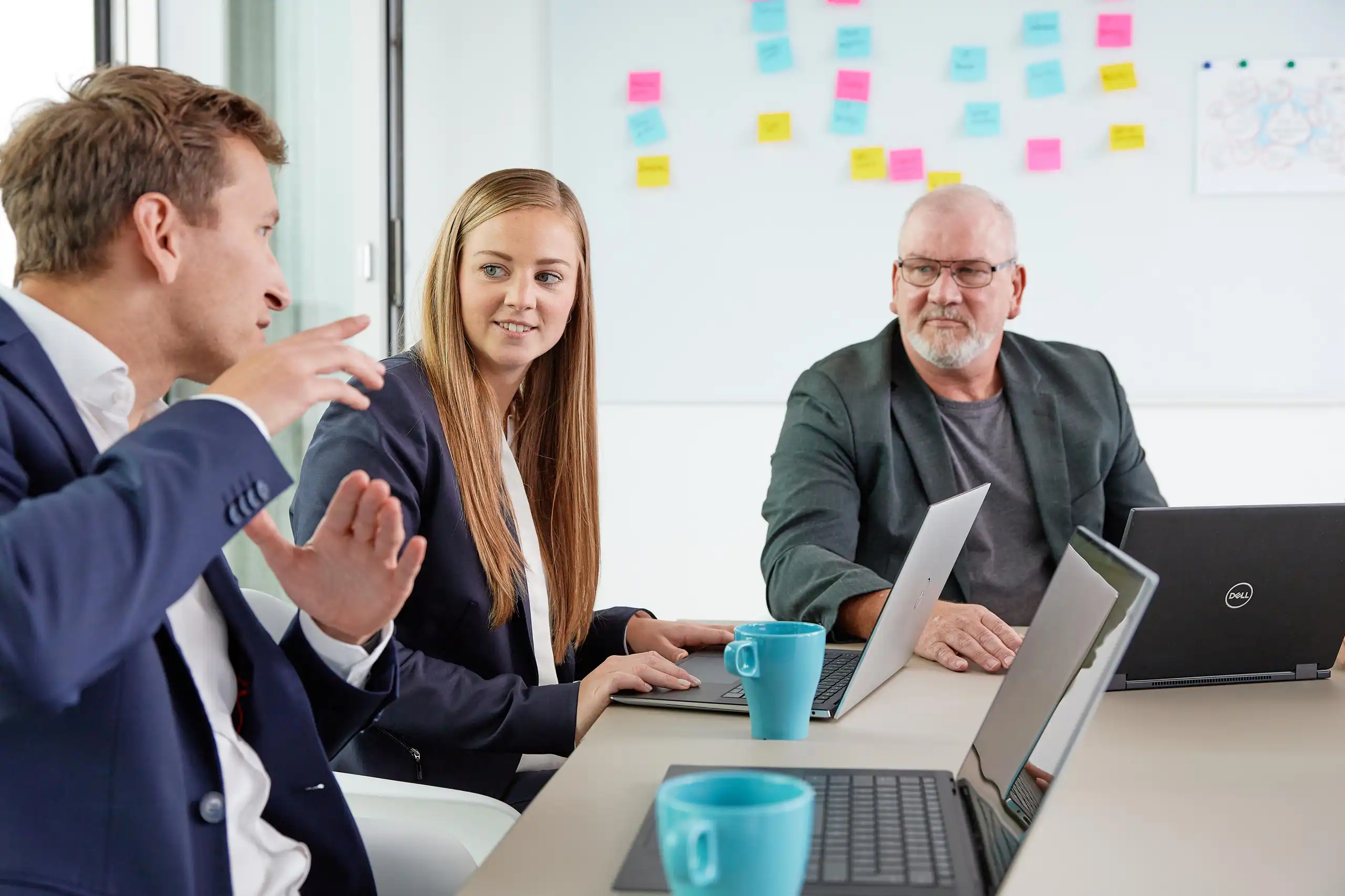 Drei Personen in Business-Kleidung sitzen mit Laptops an einem Tisch und diskutieren. Im Hintergrund ist ein Whiteboard mit bunten Haftnotizen zu sehen.