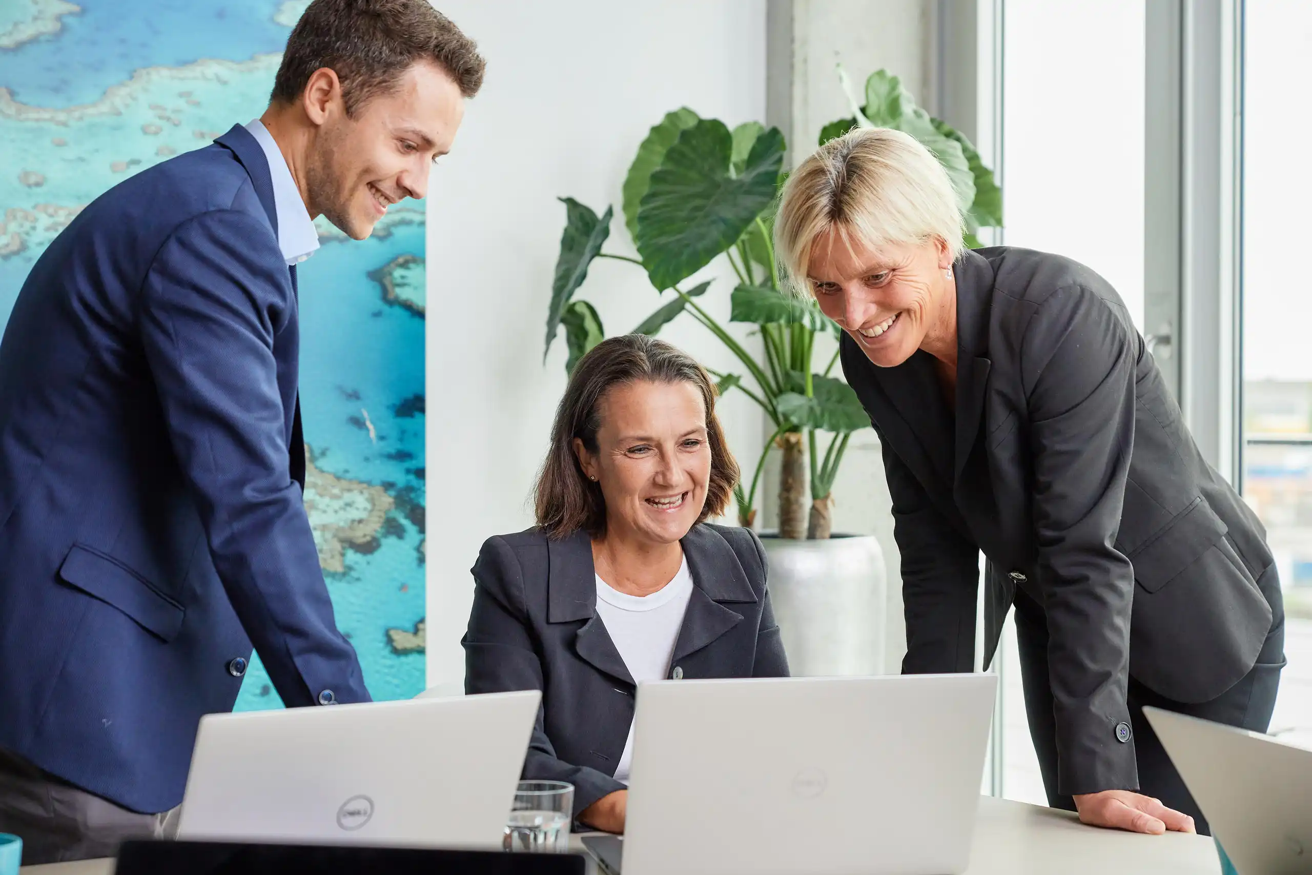 Drei Personen in Business-Kleidung stehen und sitzen an einem Tisch mit Laptops. Sie lachen und besprechen etwas in einem modernen Büro mit Pflanzen und großen Fenstern.