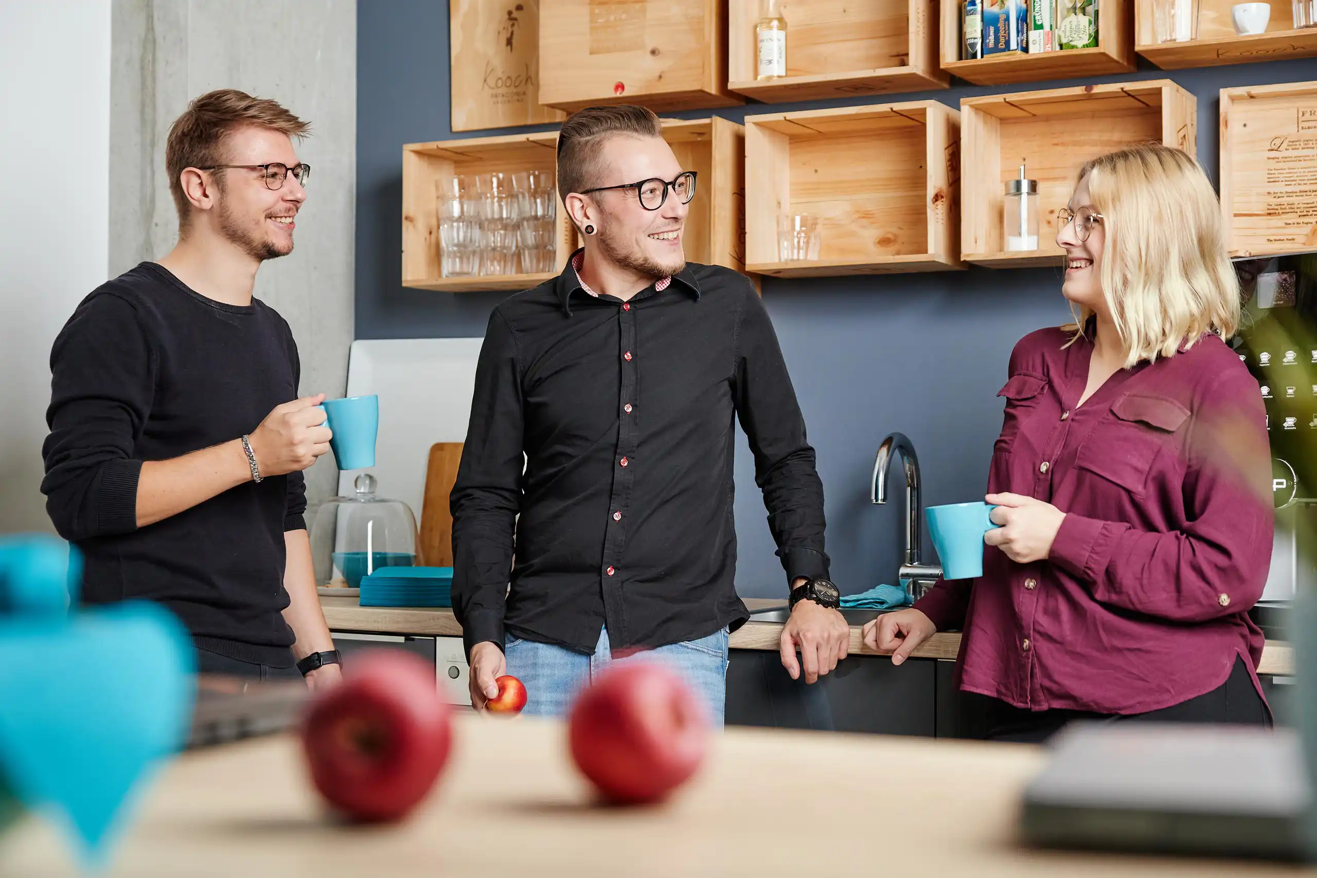 Drei Personen unterhalten sich entspannt in einer modernen Büroküche. Sie halten Tassen in der Hand und lachen.