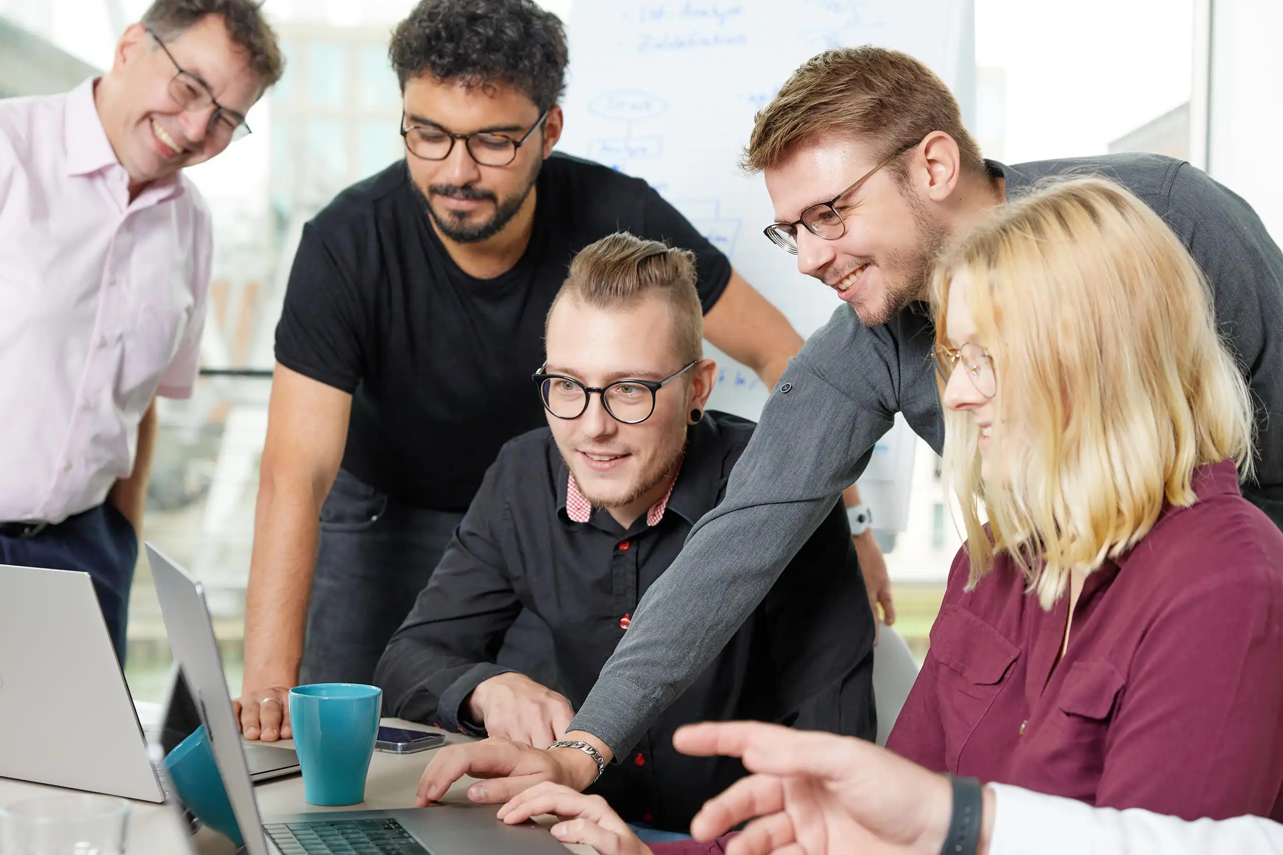 Mehrere Personen stehen und sitzen um einen Laptop, diskutieren und arbeiten zusammen. Die Stimmung ist konzentriert und kooperativ, mit einem Whiteboard im Hintergrund.