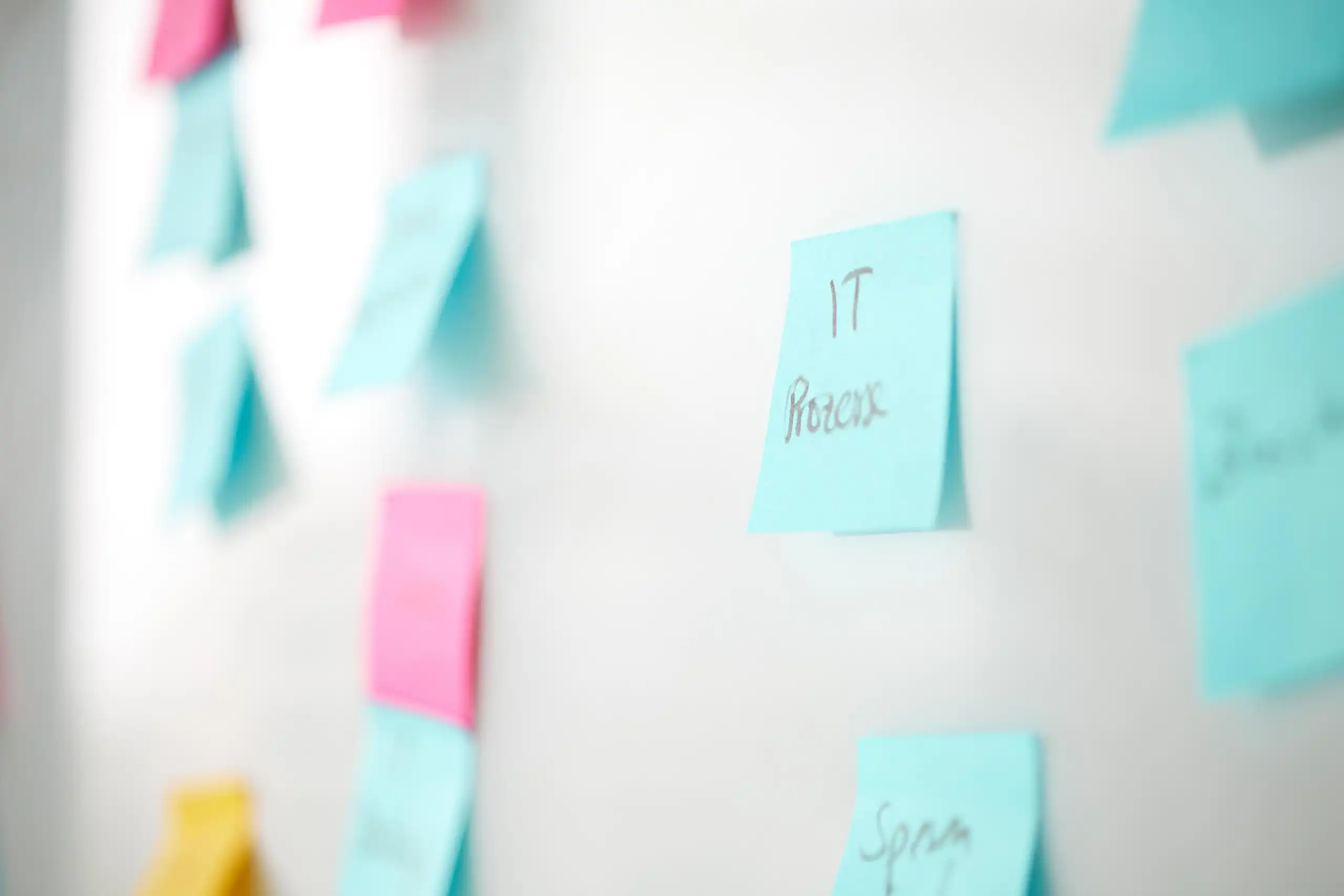 Eine weiße Pinnwand mit bunten Haftnotizen, darunter eine blaue mit der Aufschrift „IT Prozess“, die scharf im Fokus steht. Die anderen Notizen sind unscharf.