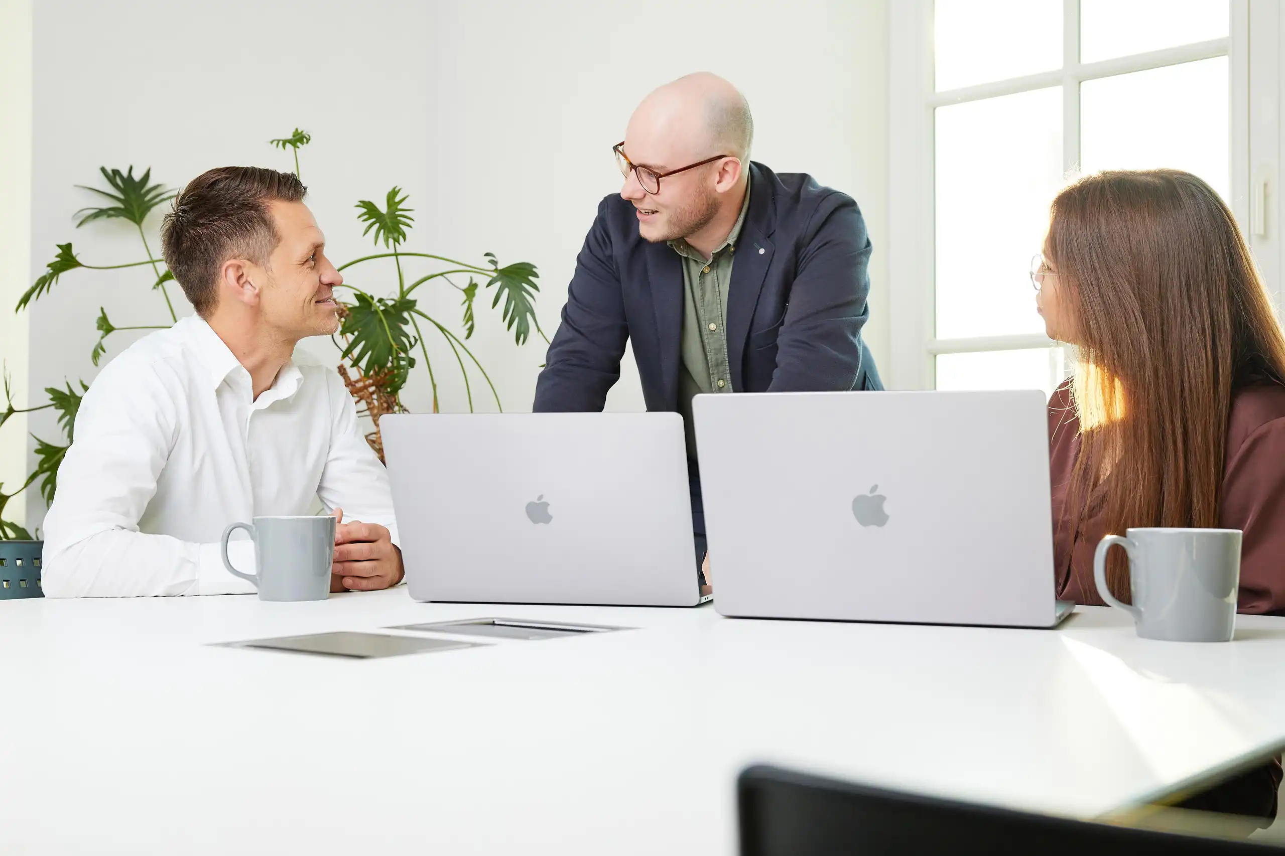 Drei Personen in Business-Kleidung besprechen sich in einem hellen Büro mit Laptops und Kaffeetassen auf dem Tisch. Eine Person steht, während die anderen sitzen.