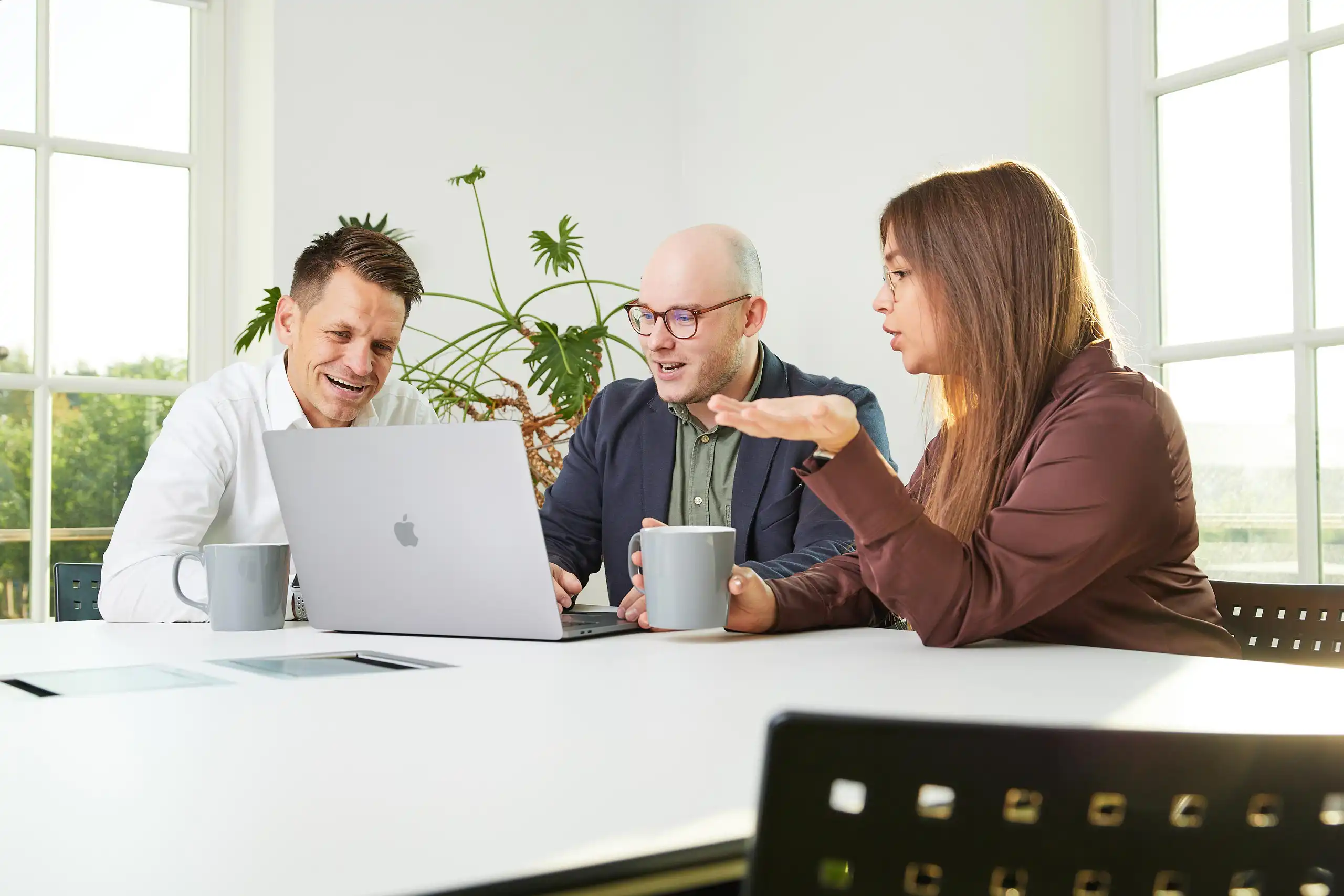 Drei Personen sitzen an einem Tisch mit Laptop und Kaffeetassen. Eine Frau gestikuliert beim Sprechen, während die beiden Männer aufmerksam zuhören und lächeln.