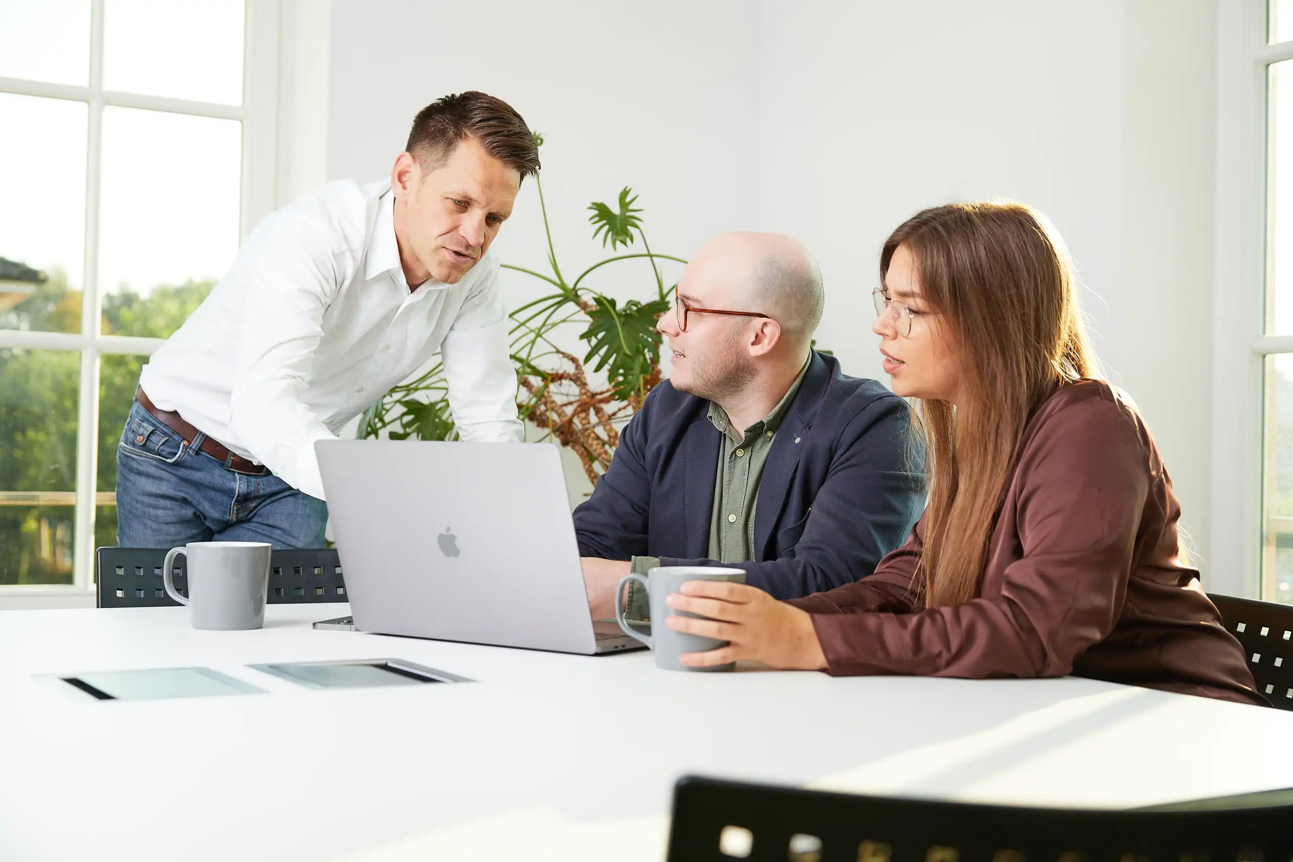 Drei Personen sitzen in einem hellen Büro an einem Tisch mit Laptop und Kaffeetassen. Ein Mann steht und erklärt etwas, während die anderen aufmerksam zuhören.