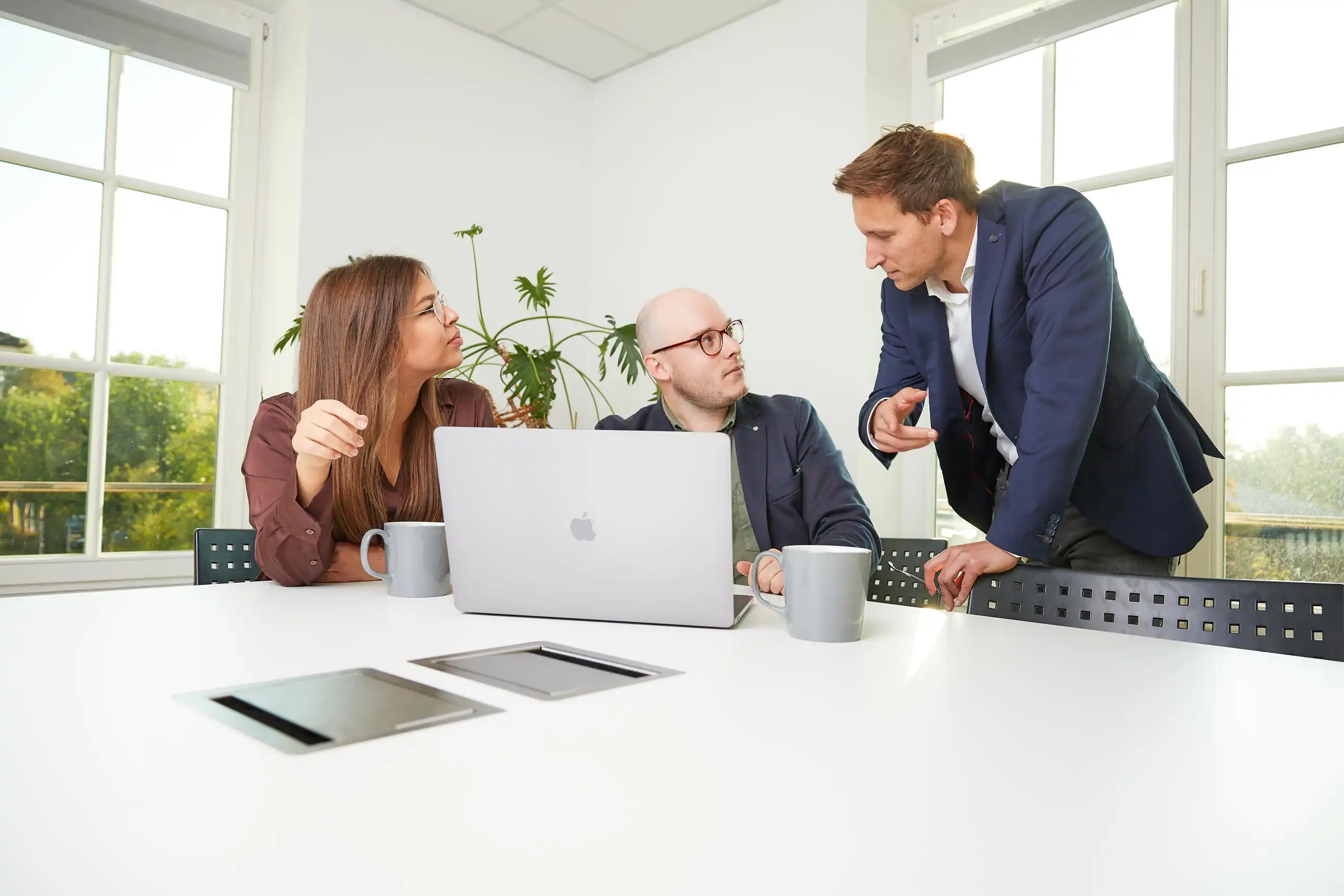 Drei Personen diskutieren an einem Tisch mit Laptop und Kaffeetassen in einem modernen, hellen Büro mit großen Fenstern und Pflanzen im Hintergrund.