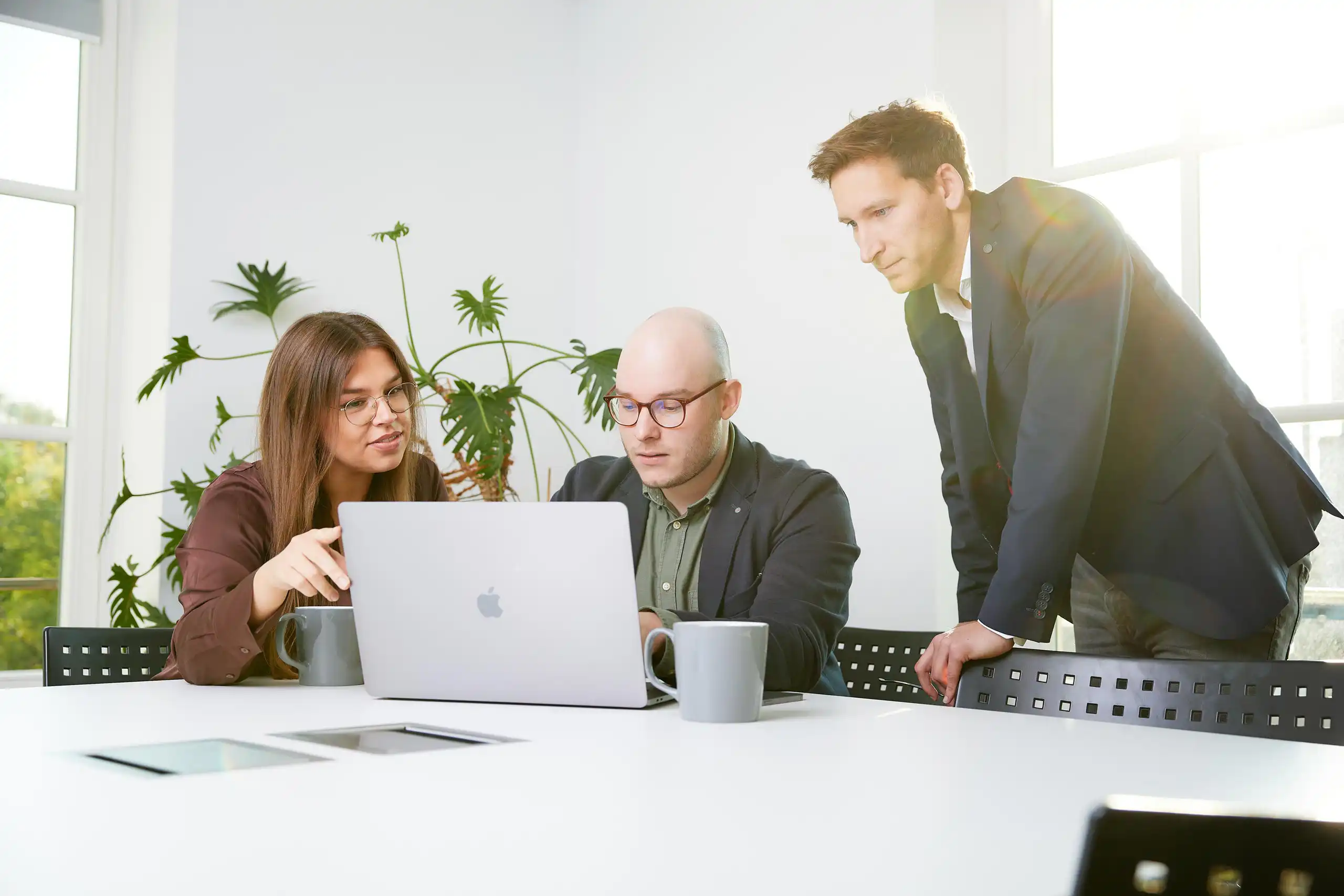 Drei Personen in Business-Kleidung sitzen an einem Tisch, schauen auf einen Laptop und diskutieren konzentriert. Sonnenlicht fällt durch große Fenster ins helle Büro.