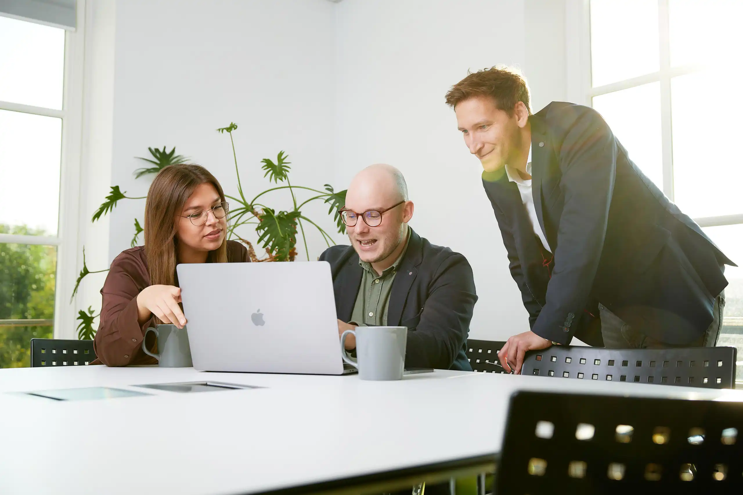 Drei Personen arbeiten gemeinsam an einem Laptop in einem hellen Büro. Eine Frau zeigt auf den Bildschirm, während die anderen lächeln und diskutieren.