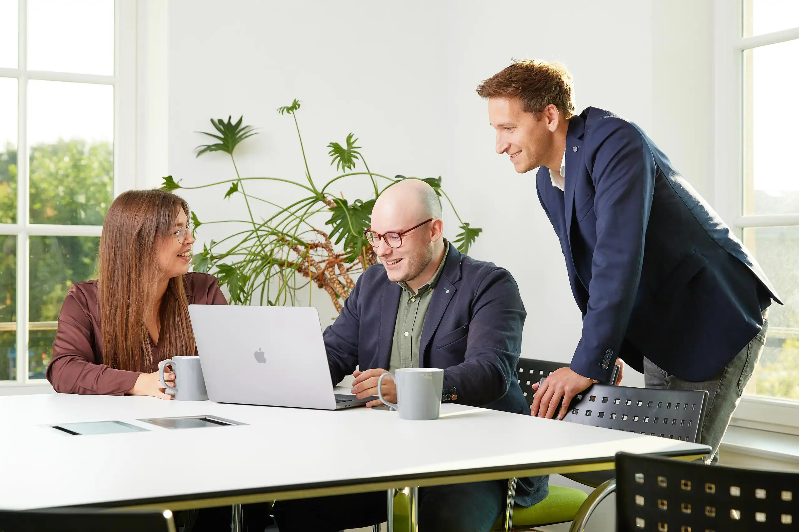 Drei Personen in Business-Kleidung sitzen an einem Tisch mit Laptops und Tassen. Sie lachen und arbeiten gemeinsam in einem modernen, hellen Büro mit Pflanzen