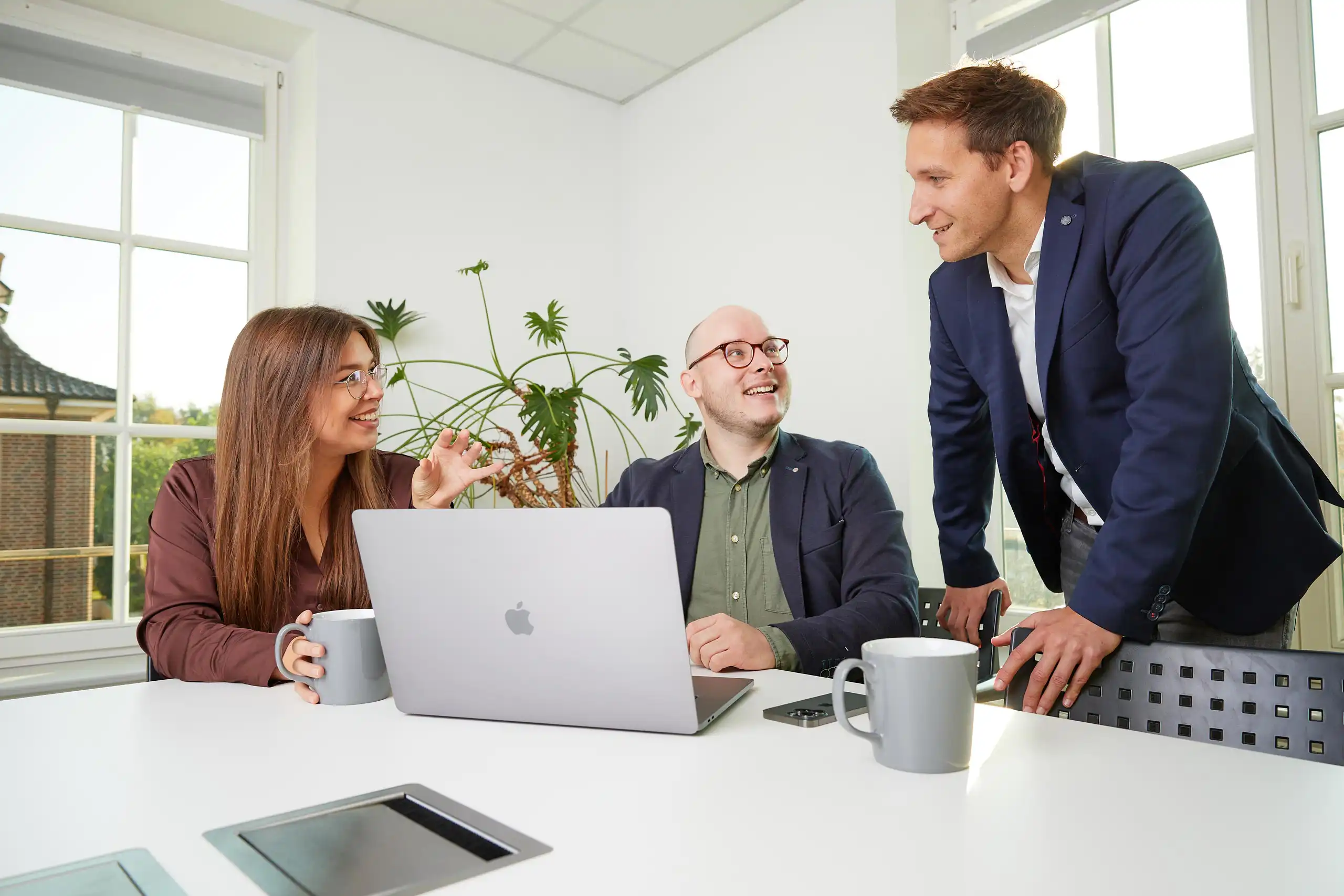 Drei Personen in Business-Kleidung sitzen und stehen um einen Tisch mit Laptops und Tassen. Sie führen eine lebhafte Diskussion in einem hellen Büro.