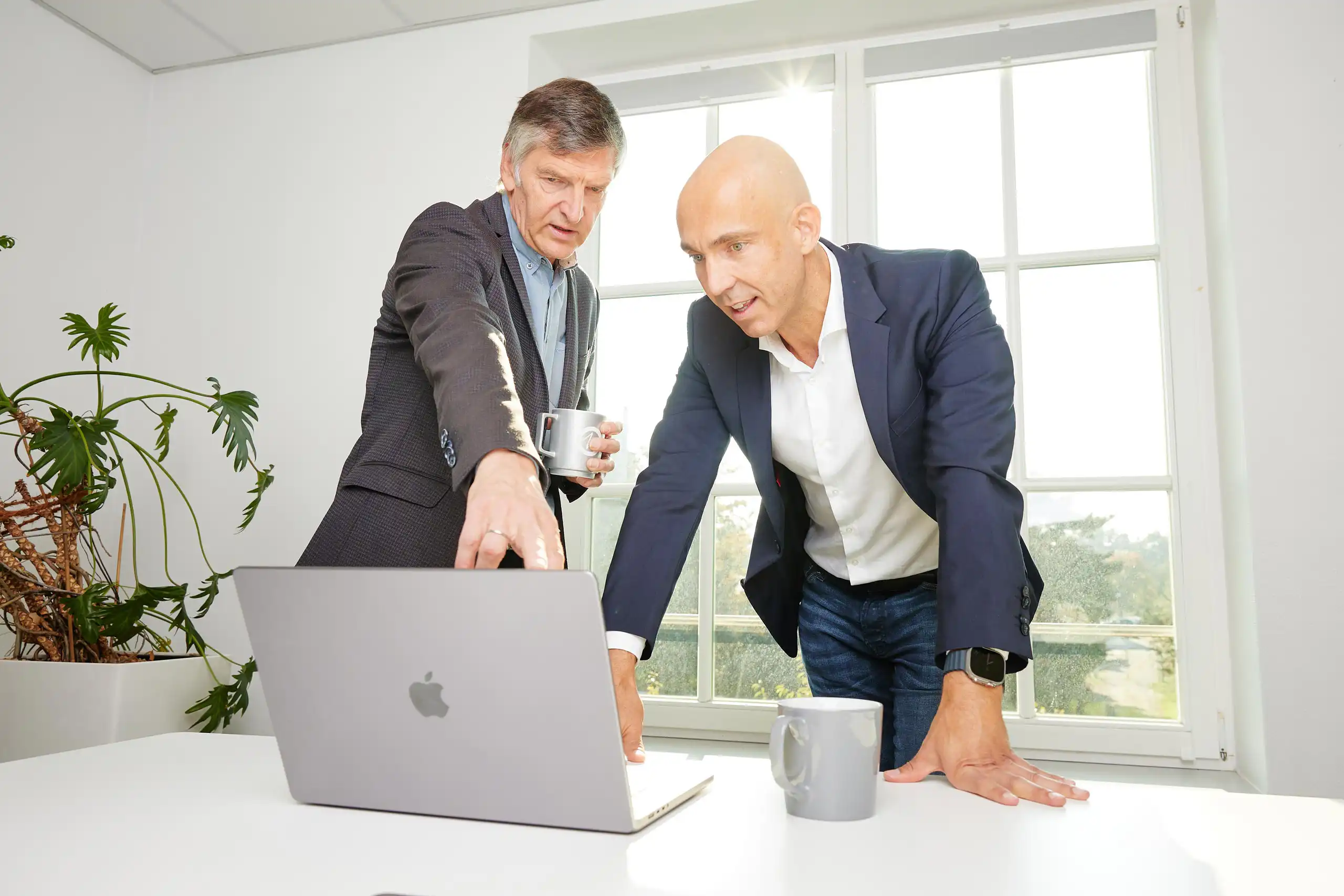 Zwei Männer in Business-Kleidung stehen an einem Tisch mit Laptop und Tassen. Einer zeigt auf den Bildschirm, während der andere sich interessiert nach vorne lehnt.