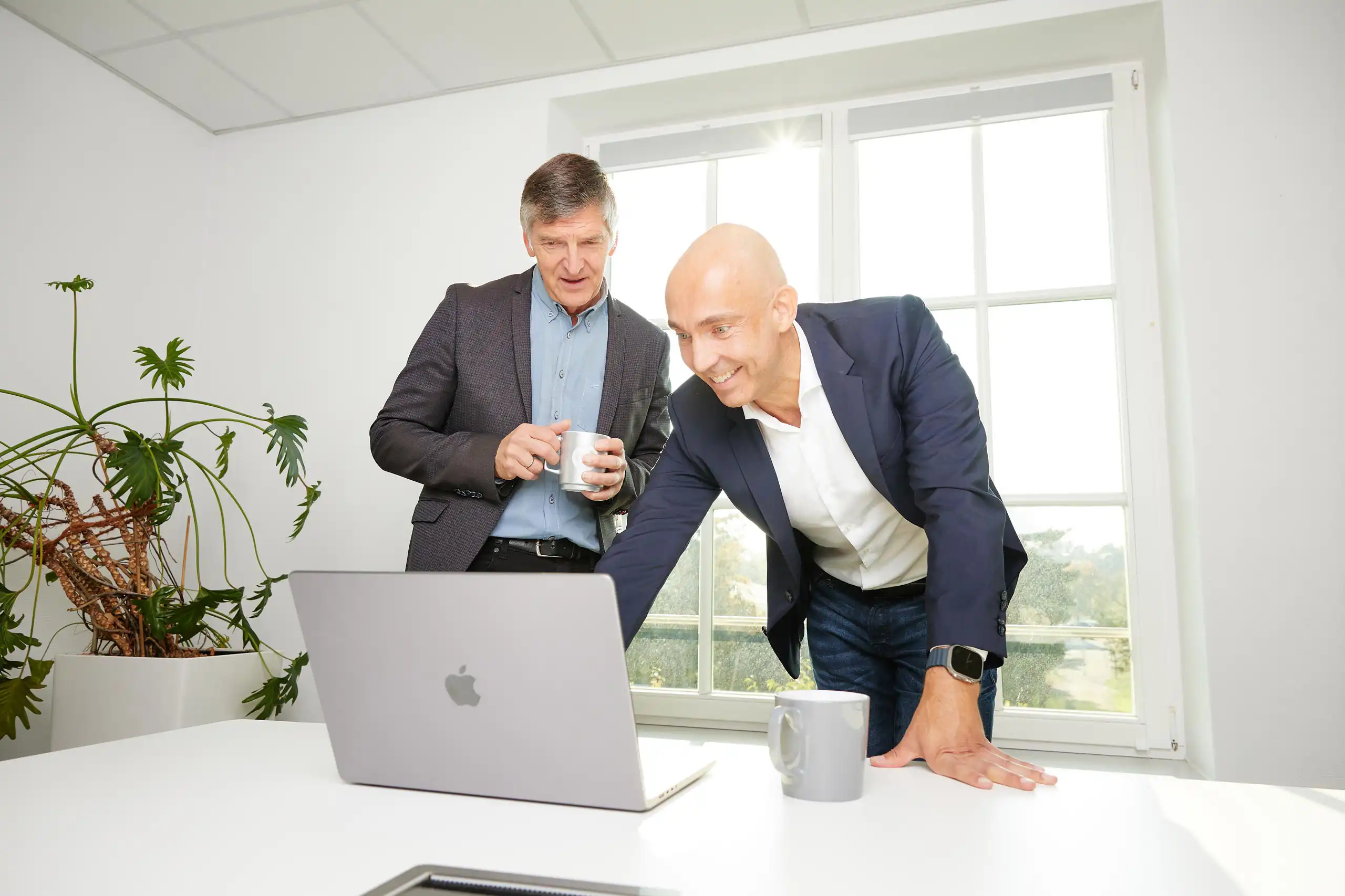 Zwei Männer in Business-Kleidung stehen an einem Tisch mit einem Laptop. Einer zeigt lächelnd auf den Bildschirm, während der andere zuschaut.