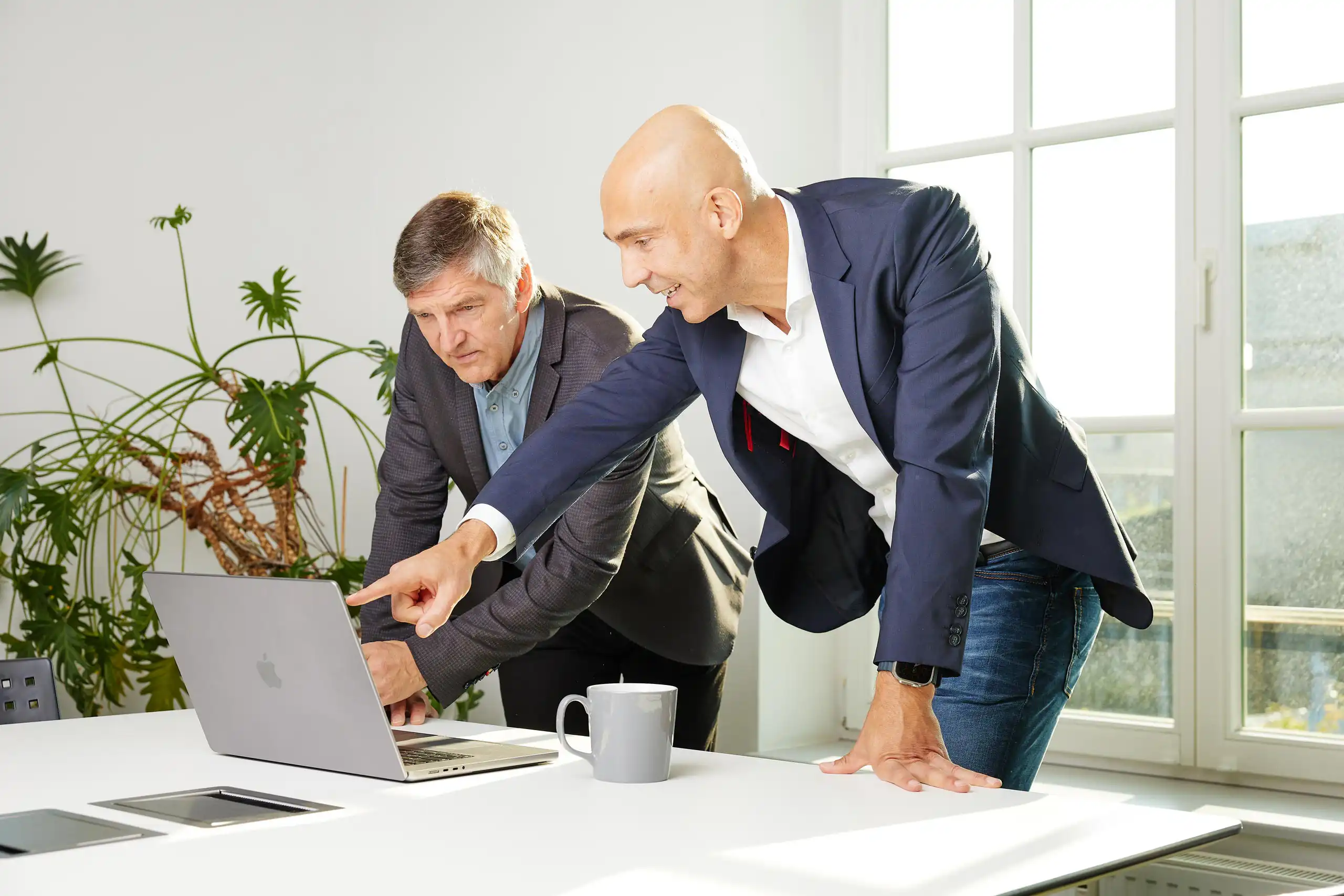 Zwei Männer in Business-Kleidung beugen sich über einen Laptop. Einer zeigt auf den Bildschirm, während der andere konzentriert schaut.