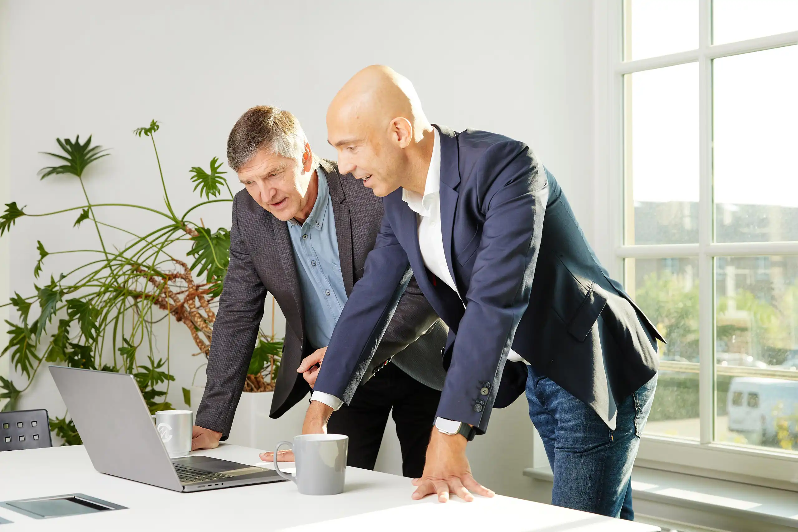 Das Bild zeigt zwei Männer in Business-Kleidung, die sich über einen Laptop beugen und gemeinsam arbeiten.