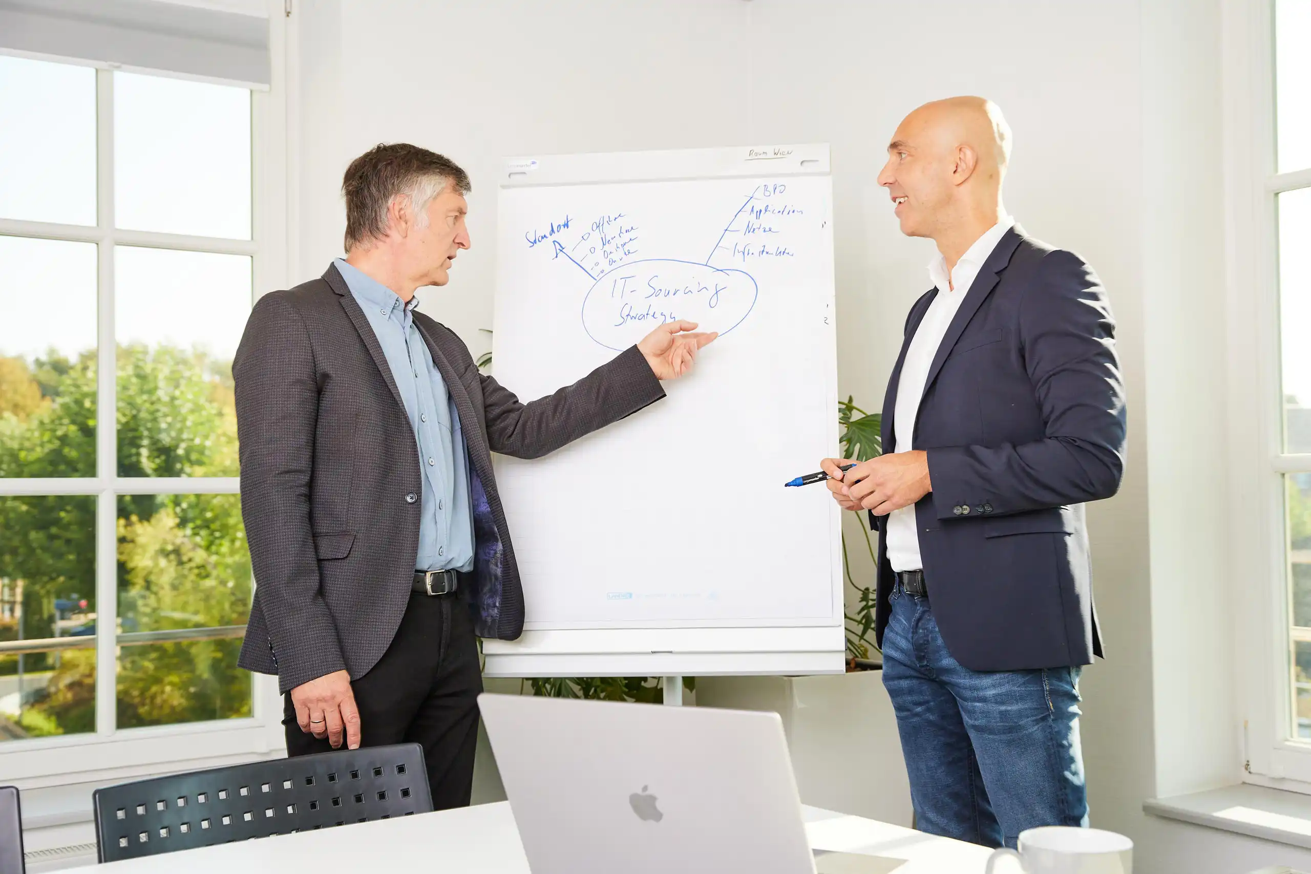 Zwei Männer in Business-Kleidung stehen vor einem Flipchart und besprechen eine IT-Sourcing-Strategie. Einer zeigt auf die Skizze, der andere hält einen Stift.