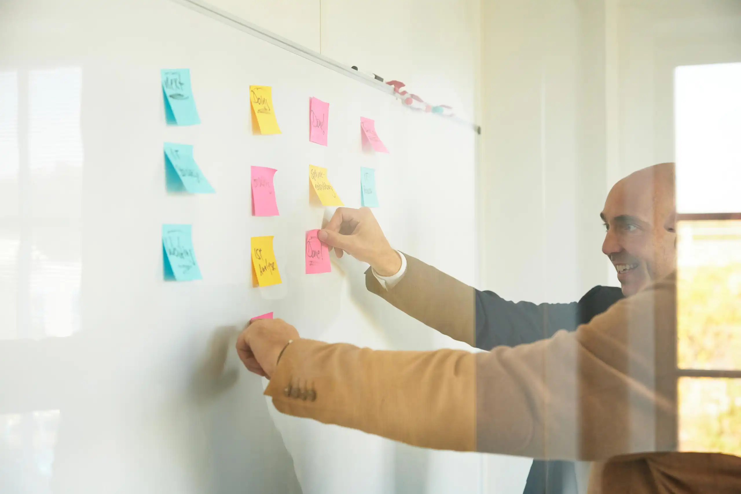 Zwei Männer kleben bunte Haftnotizen auf ein Whiteboard und arbeiten gemeinsam an Ideen.