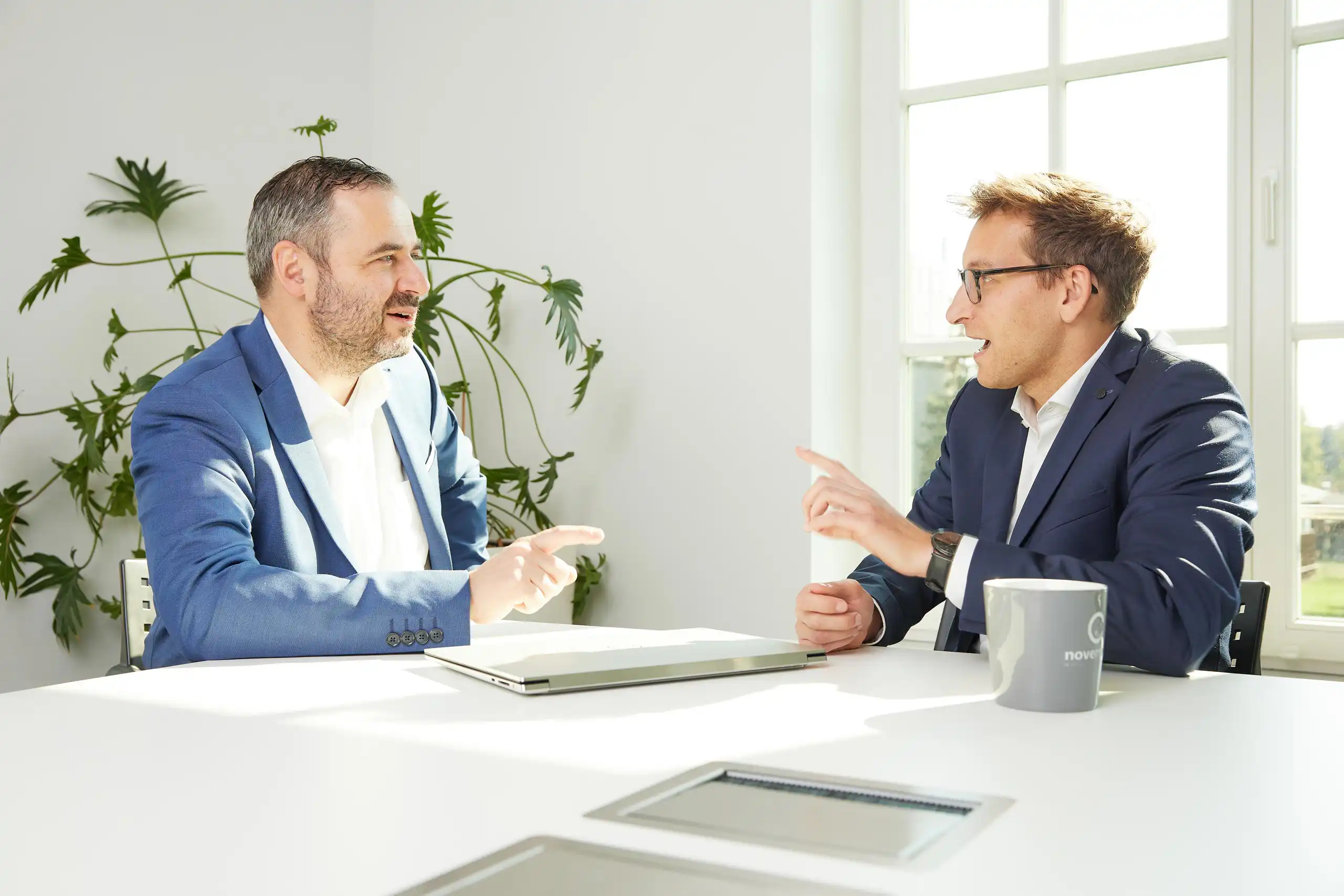 Zwei Männer in Business-Kleidung unterhalten sich angeregt in einem sonnigen Büro. Ein Laptop und eine Tasse stehen auf dem Tisch.