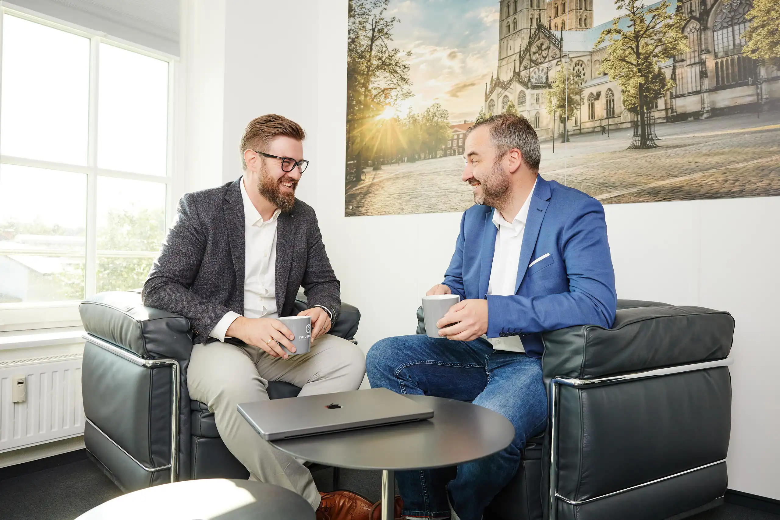 Zwei Männer sitzen entspannt in Sesseln, halten Kaffeetassen und unterhalten sich lachend. Auf dem Tisch liegt ein Laptop, im Hintergrund ein Stadtbild mit Kirche.