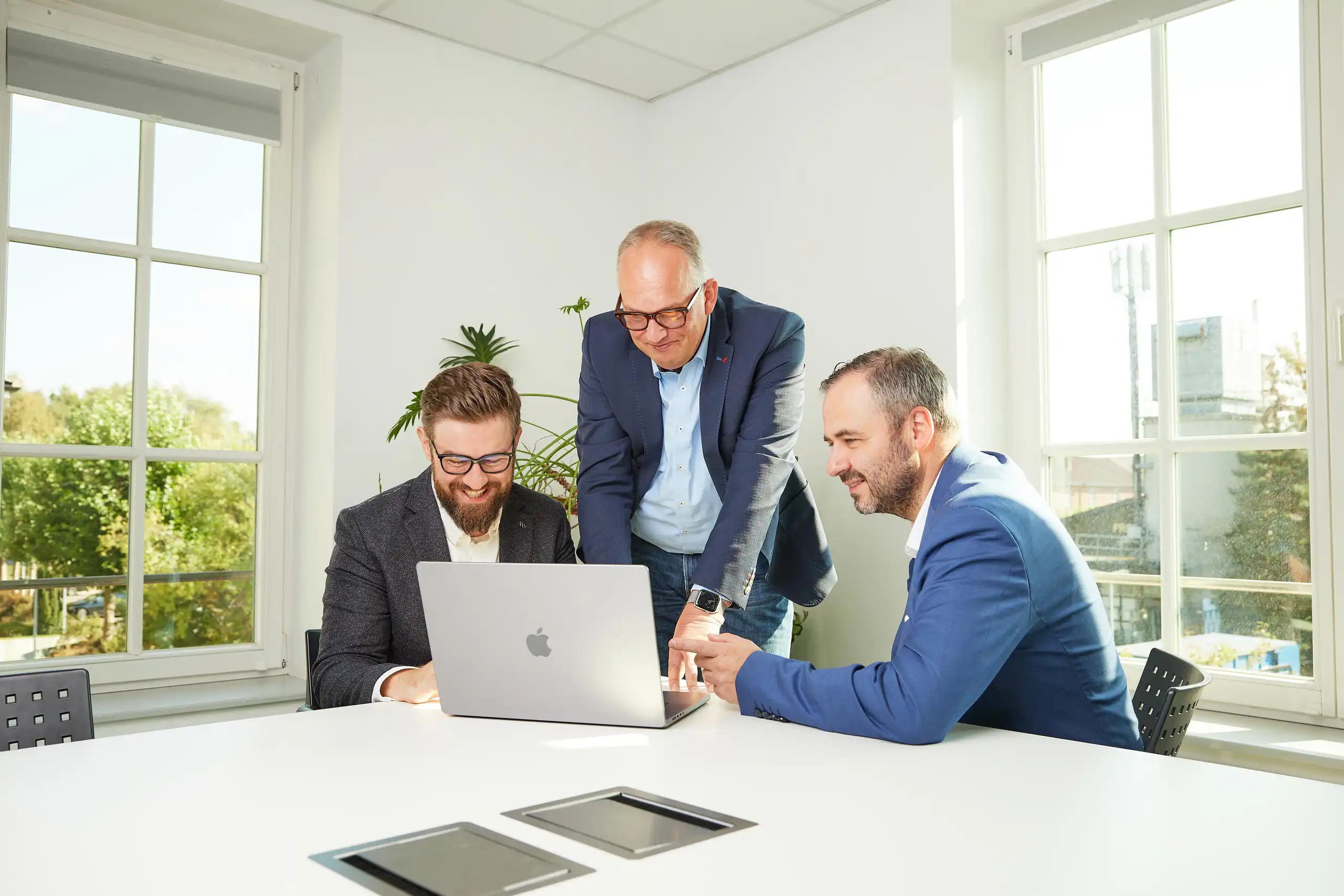 Drei Männer in Business-Kleidung arbeiten gemeinsam an einem Laptop in einem hellen Büro. Sie lachen und diskutieren, während sie auf den Bildschirm schauen.