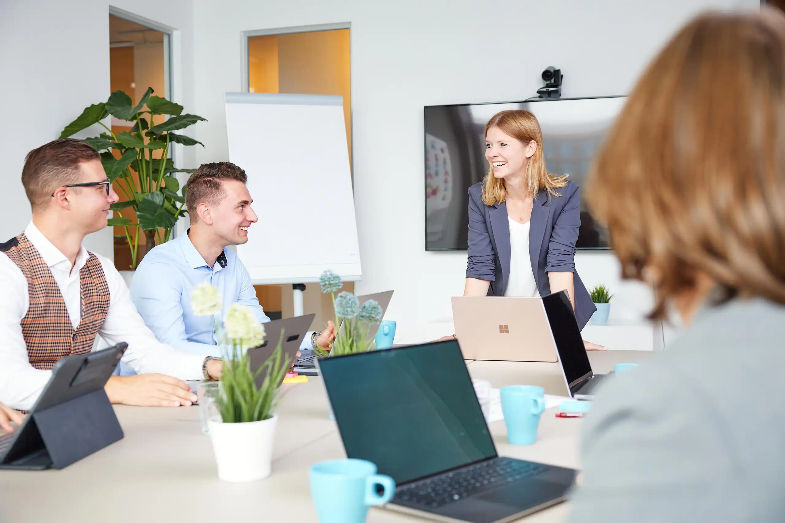 Eine Frau steht lächelnd an einem Konferenztisch, spricht mit Kollegen, die an Laptops arbeiten. Helle, moderne Büroumgebung mit Pflanzen und Flipchart im Hintergrund.
