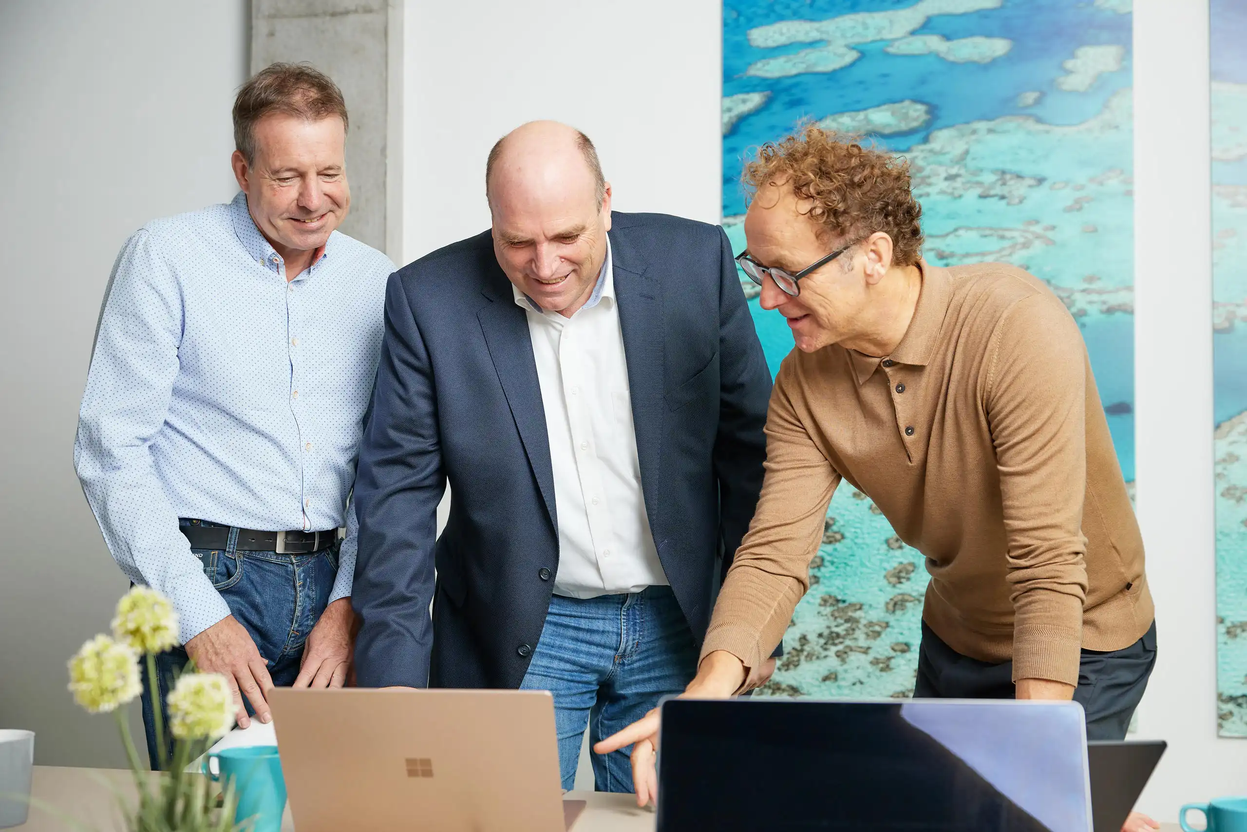 Drei Männer stehen an einem Tisch und schauen gemeinsam auf einen Laptop. Sie lachen und diskutieren angeregt in einer modernen Büroatmosphäre.