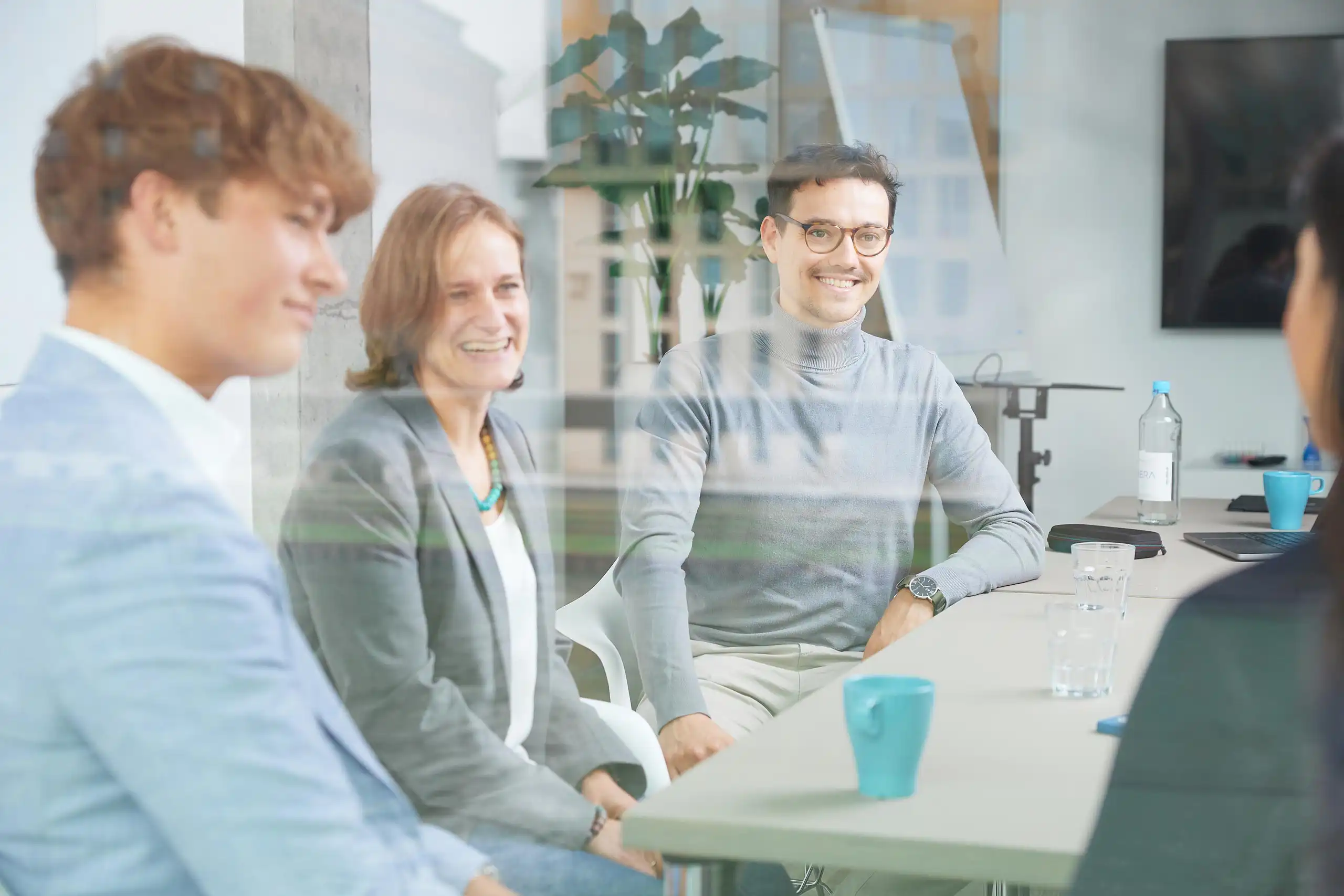 Vier Personen sitzen in einem modernen Besprechungsraum und führen ein freundliches Gespräch. Sie lachen und wirken entspannt. Das Bild ist durch eine Glasscheibe aufgenommen.