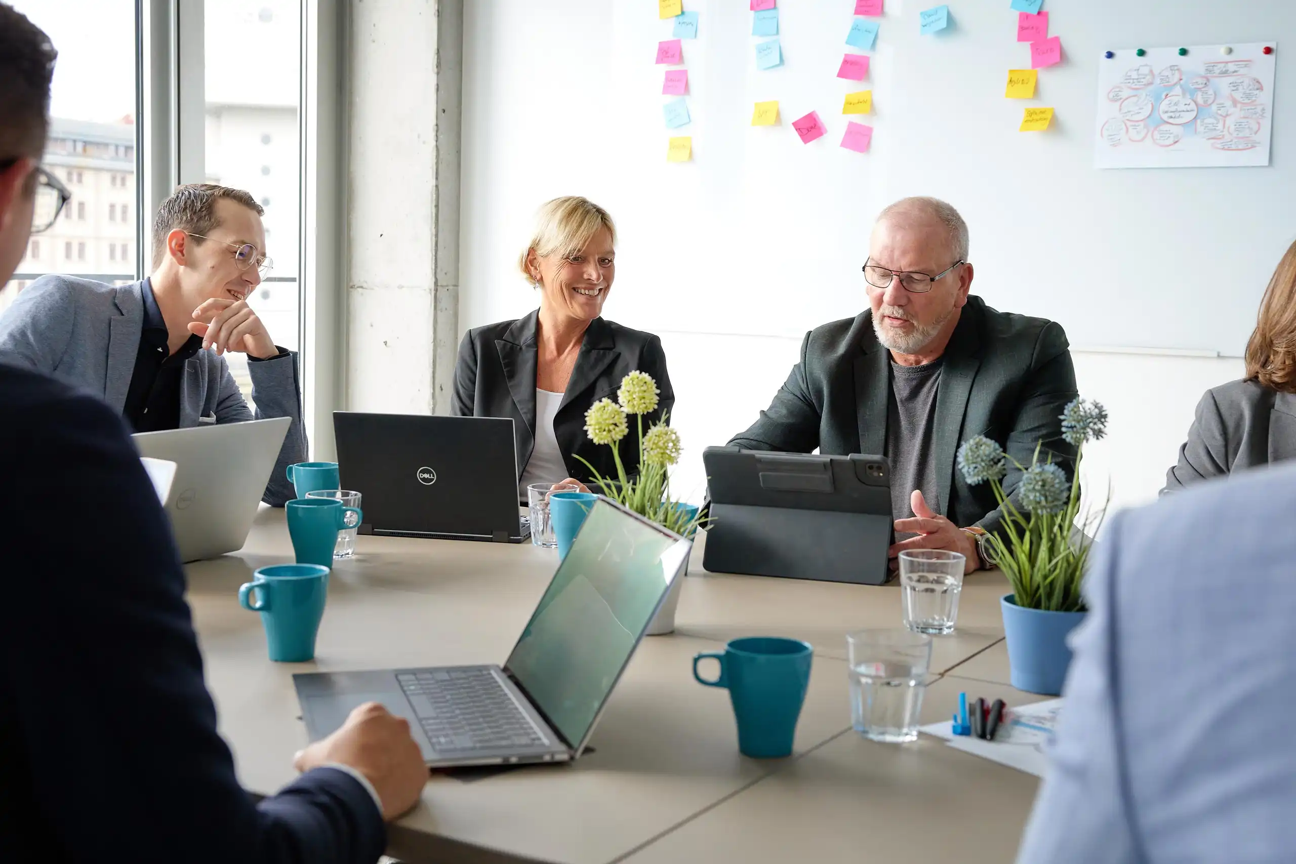 Fünf Personen in Business-Kleidung sitzen in einem modernen Büro an einem Tisch mit Laptops, Tablets und Notizen, während sie eine Besprechung führen.