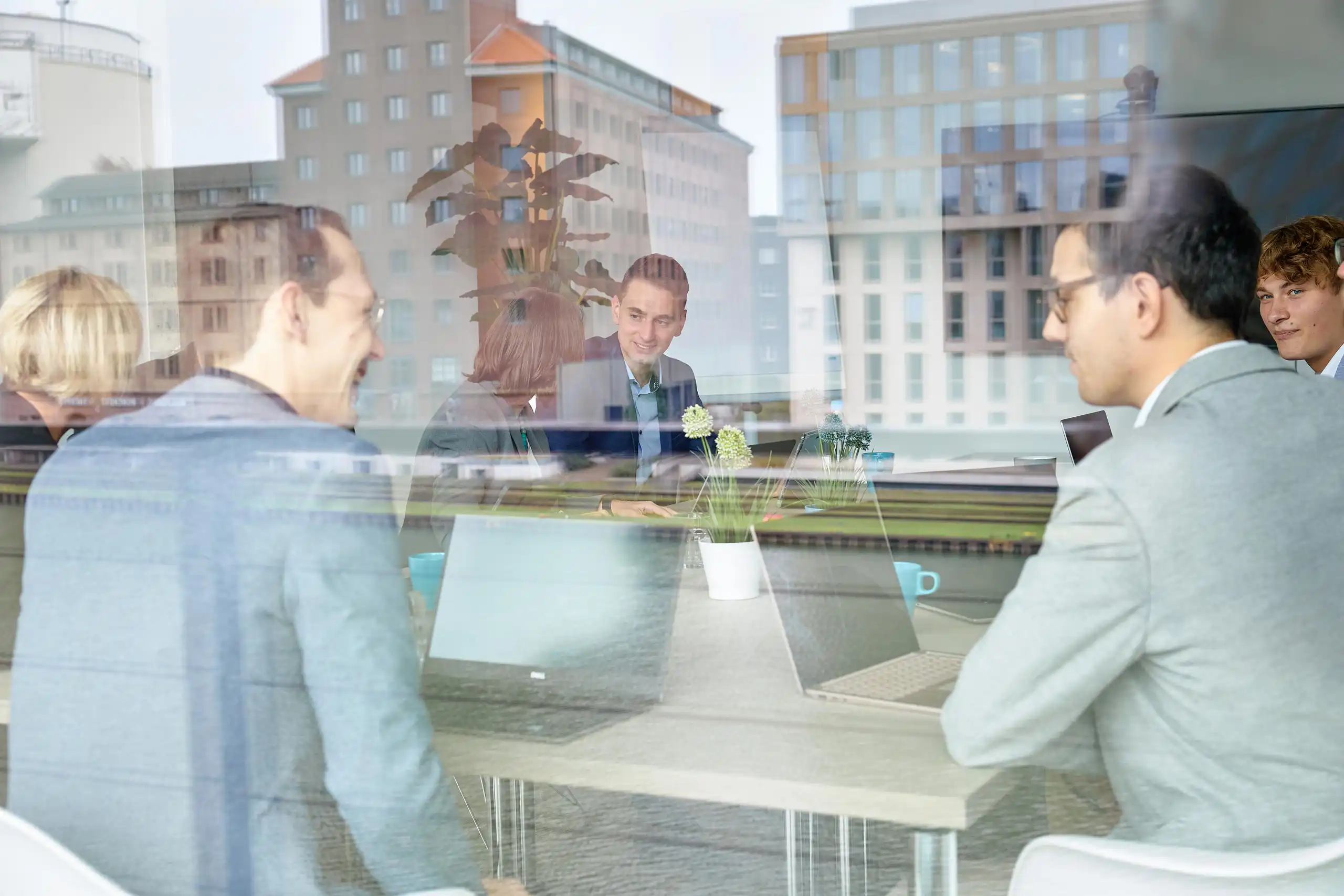 Mehrere Personen sitzen in einem modernen Büro an einem Tisch mit Laptops und Pflanzen. Die Spiegelung im Fenster zeigt die Stadtlandschaft im Hintergrund.