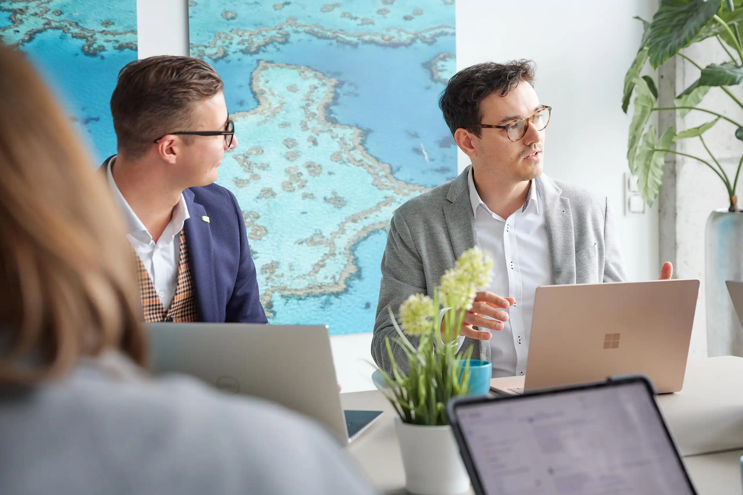 Mehrere Personen sitzen in einem modernen Büro an einem Tisch mit Laptops und Pflanzen. Eine Person spricht gestikulierend, während die anderen zuhören.