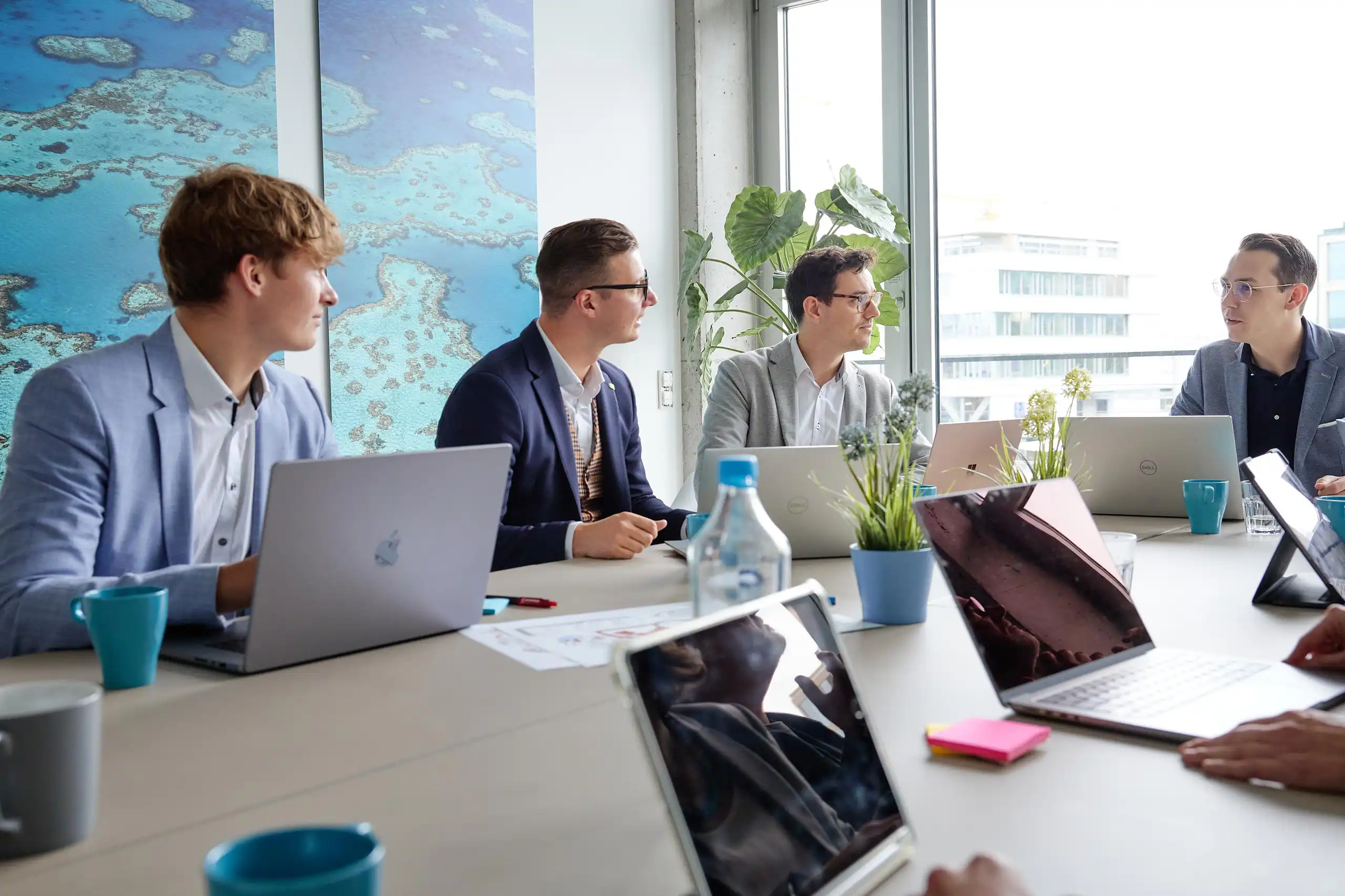 Ein Team in Business-Outfits sitzt in einem modernen Konferenzraum mit Laptops, Notizen und Pflanzen auf dem Tisch.