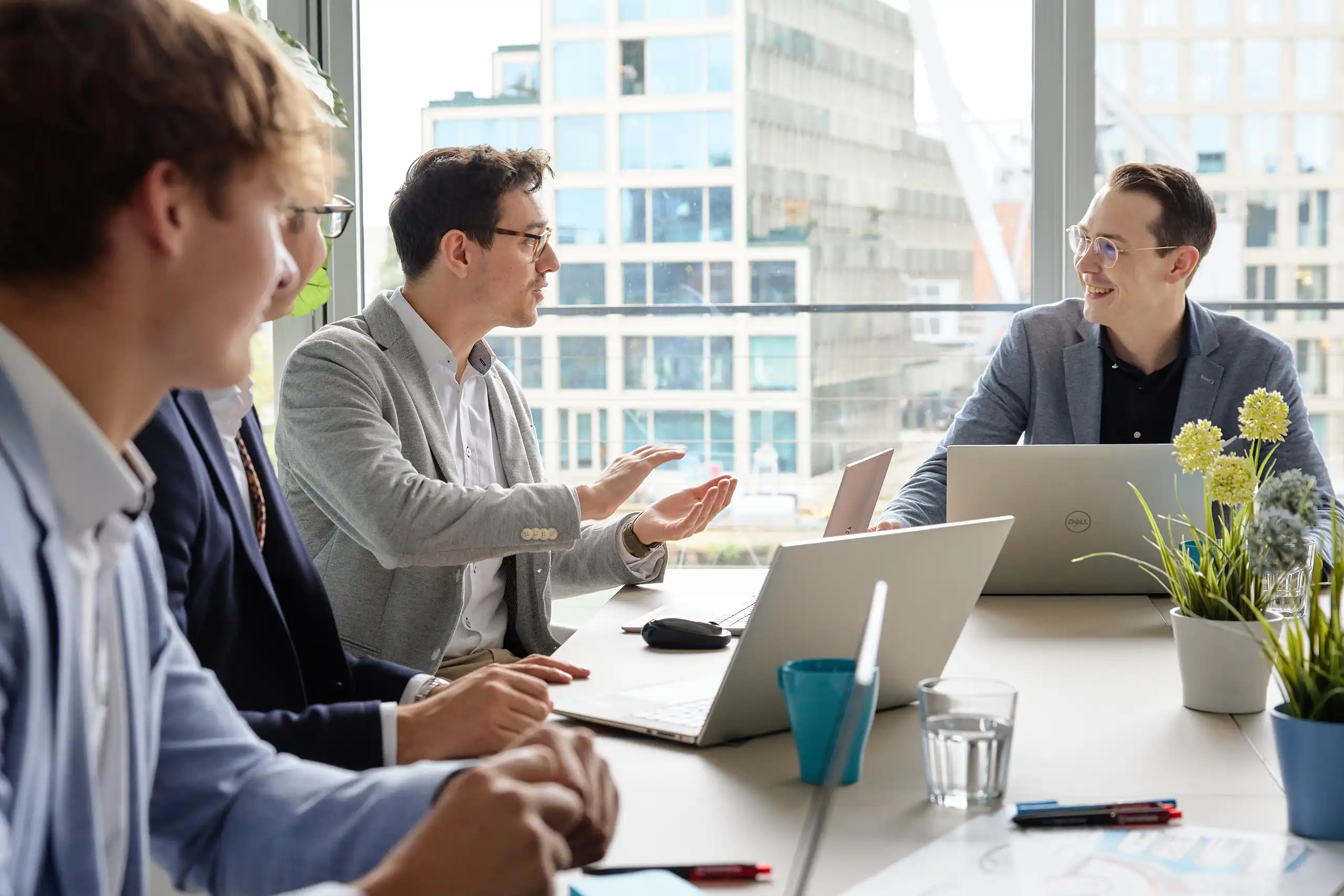 Mehrere Männer in Business-Kleidung sitzen an einem Tisch mit Laptops und Pflanzen. Einer spricht gestikulierend, während die anderen zuhören und lächeln.