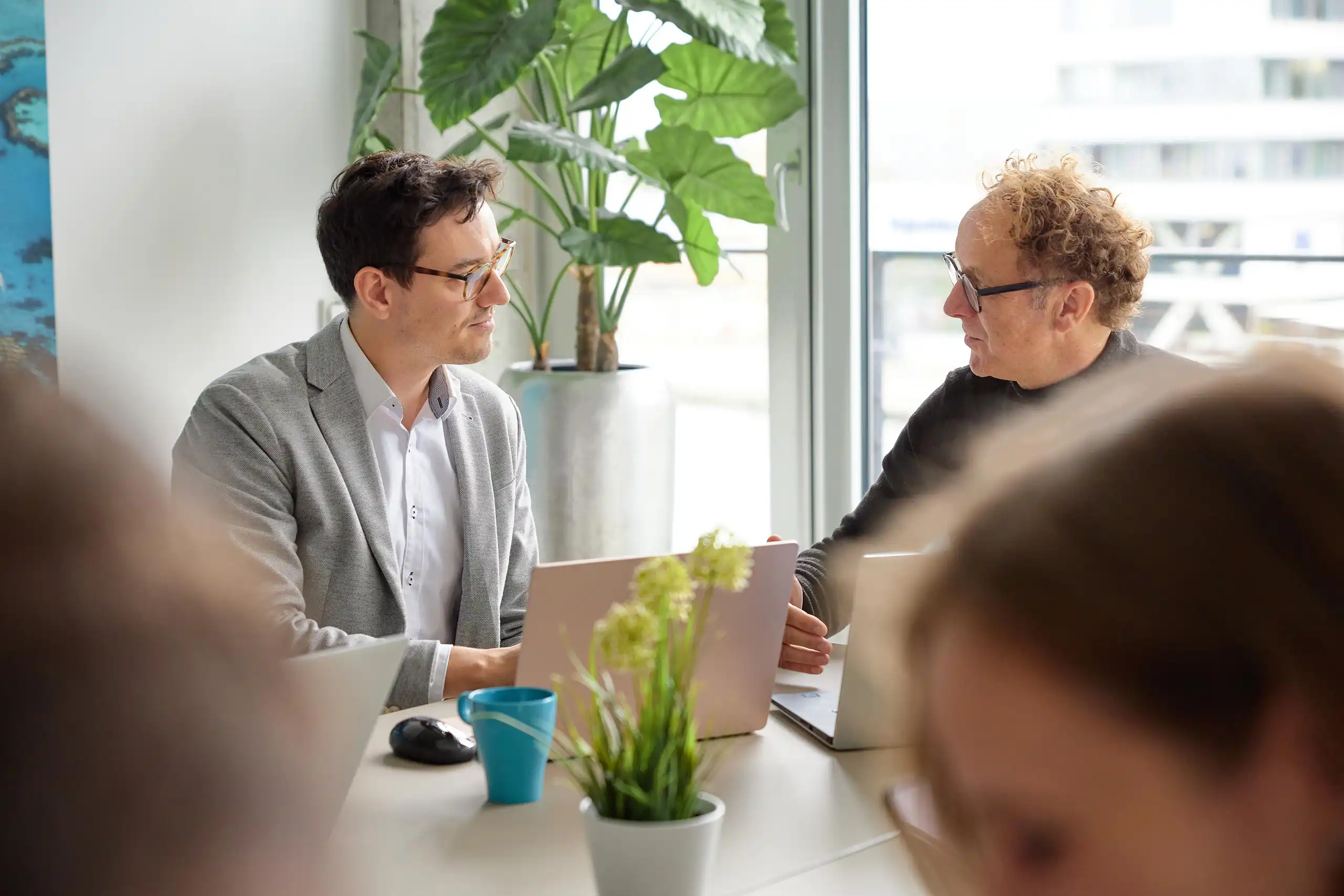 Zwei Männer sitzen in einem modernen Büro mit Laptops und Pflanzen, vertieft in ein Gespräch. Im Vordergrund sind unscharf weitere Personen erkennbar.