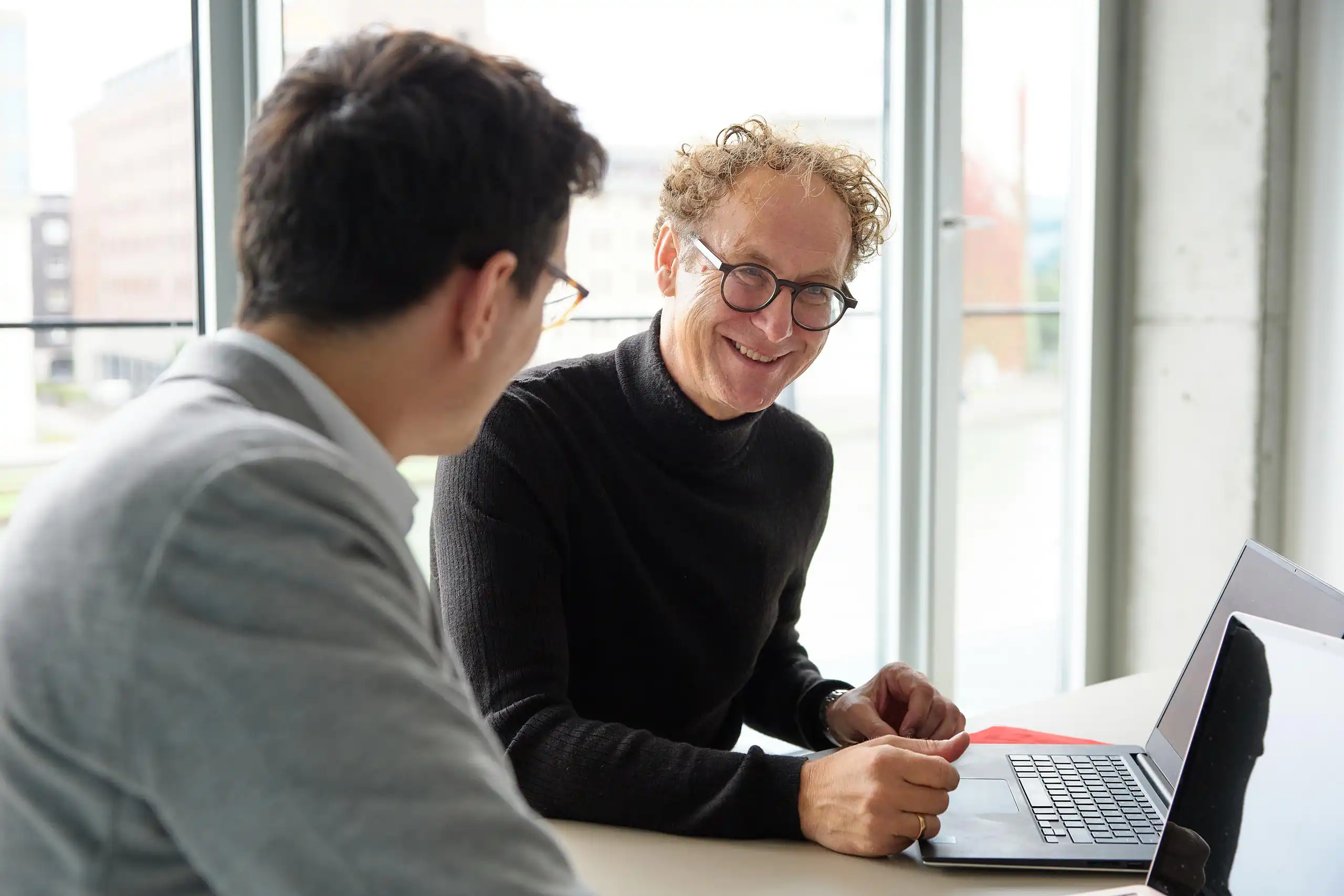Zwei Männer sitzen an einem Tisch mit Laptops und unterhalten sich. Einer lächelt freundlich, während sie sich in einem modernen Büro mit großen Fenstern austauschen.