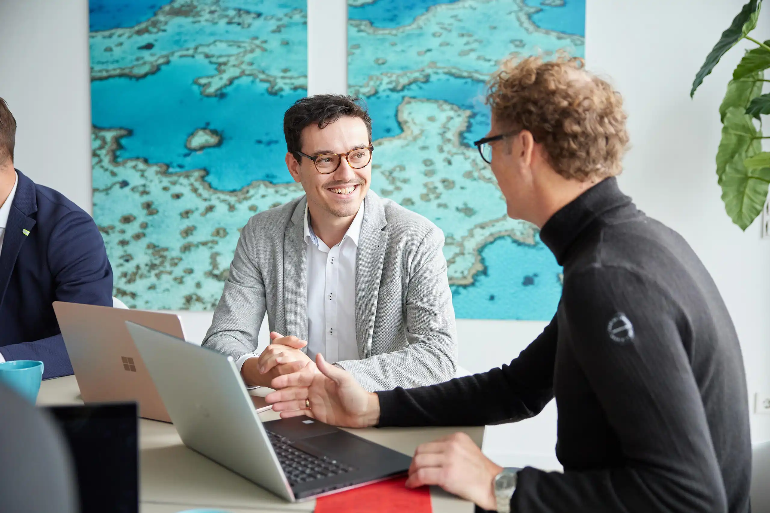 Zwei Männer in Business-Kleidung führen ein Gespräch in einem modernen Büro. Beide lächeln, während sie sich austauschen und an Laptops arbeiten.